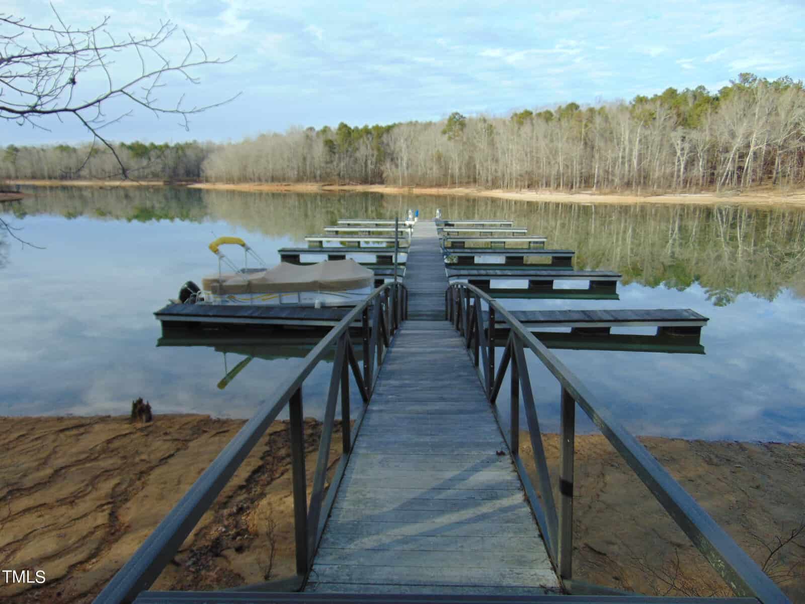 A serene lake view featuring a narrow metal pier leading to several empty boat docks, surrounded by a calm water body mirror-reflecting clear blue skies and bare trees.