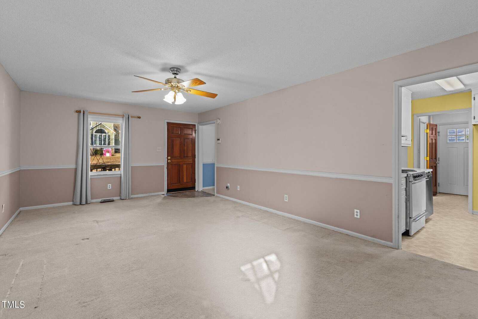 A vacant room with beige walls, carpet flooring, a ceiling fan, and a door leading outside.