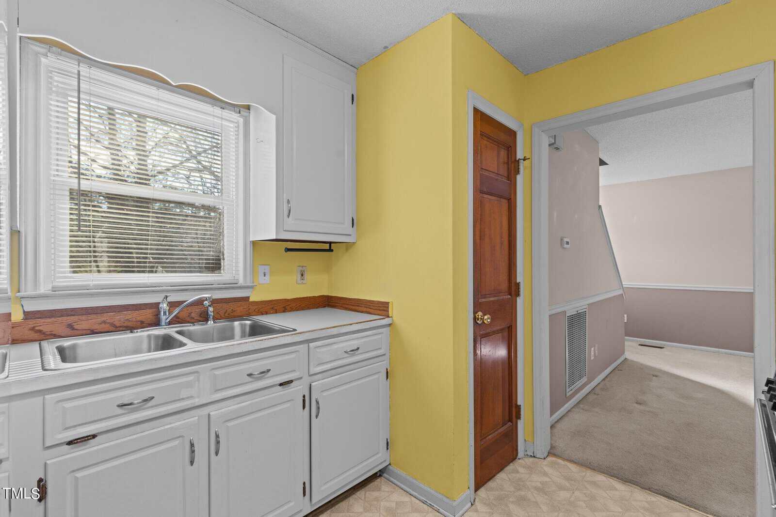 This image shows a corner of a kitchen with white cabinetry and a stainless steel sink next to a yellow wall, with a view into an adjoining room painted pink.
