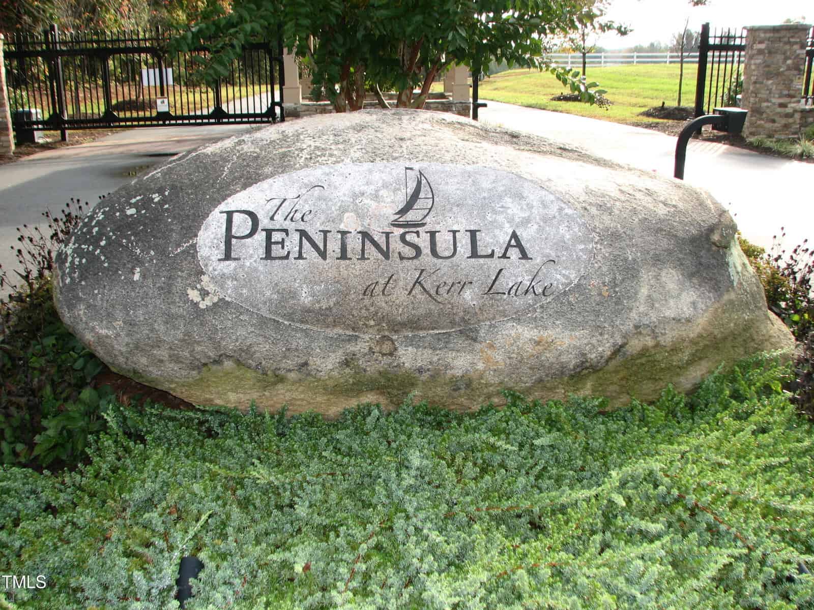 A large boulder engraved with "23 Waterstone Lane, Henderson NC" surrounded by low-growing green plants, positioned near a metal gate entrance.