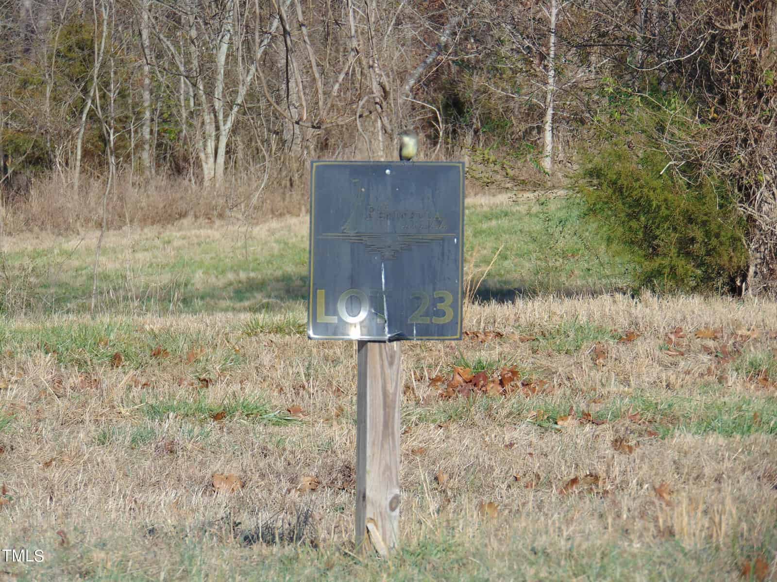 A wooden post holds up a weathered, metal real estate sign reading "lot 23" in a grassy field, with trees and a clear sky in the background.
