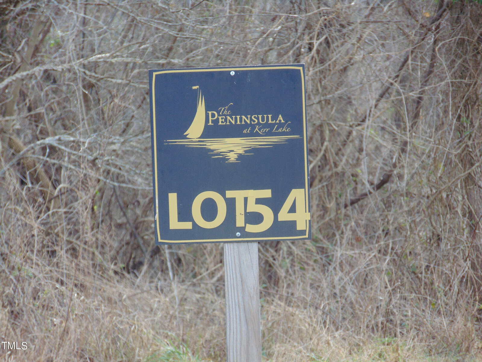 A sign indicating "lot 54" at "the peninsula at kerr lake," with a logo of a sailboat above the text, mounted on a post with vegetation in the background.