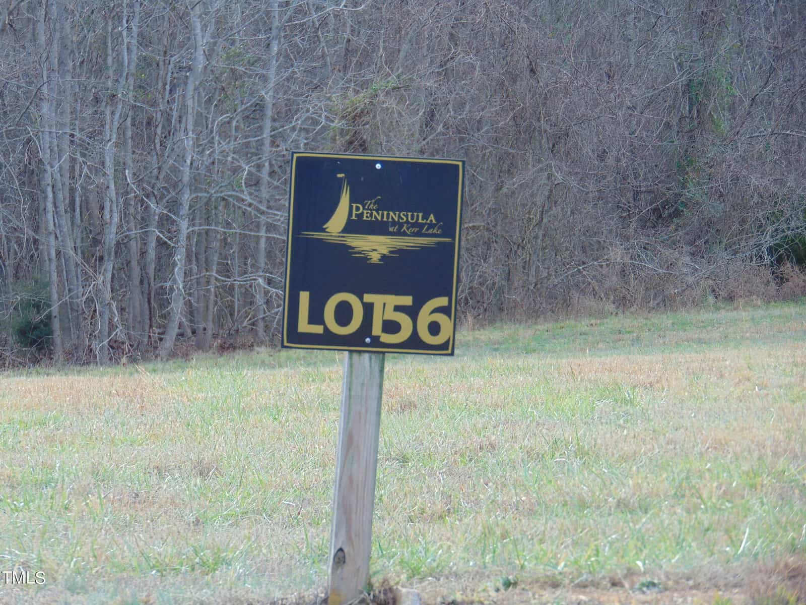 A sign reading "peninsula at the lake, lot 56" stands in a grassy field with trees in the background. the sign is black with gold lettering and an emblem depicting a stylized sun over waves.