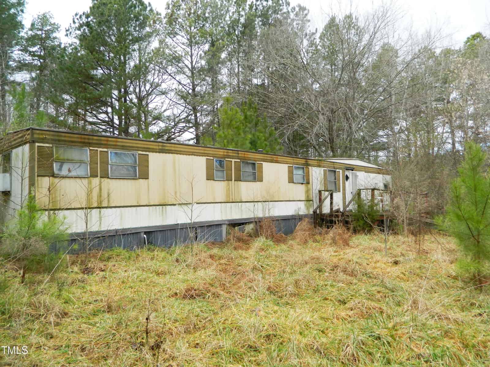 An old and weathered mobile home surrounded by overgrown grass and shrubs, set in a wooded area with tall trees in the background under a cloudy sky.