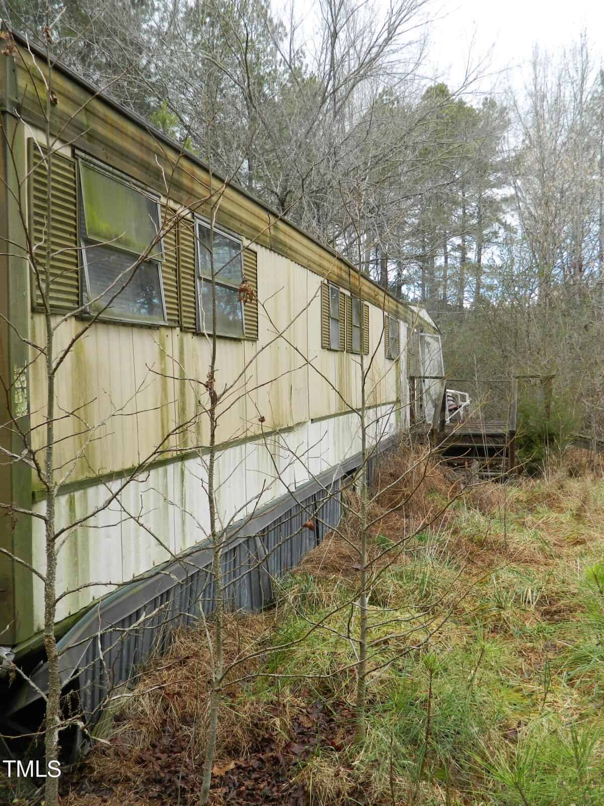 An old, weathered trailer in a grassy, overgrown area with a wooden porch. The exterior is faded with moss and dirt, and tall trees stand behind it under a cloudy sky.