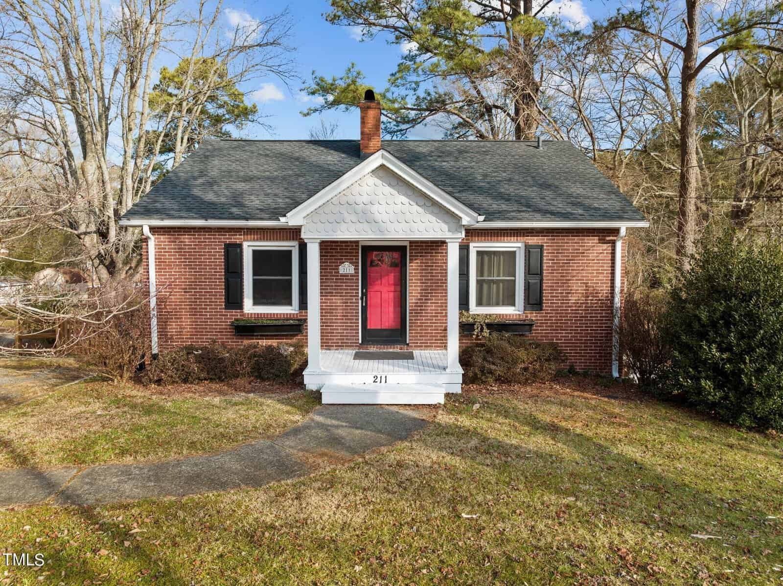 Nestled on 211 Park Avenue, this small, charming brick house with a gabled roof and a red front door captures the essence of Creedmoor. Surrounded by trees, it features white trim, black shutters, and a concrete path leading to the entrance. The grassy lawn is complemented by shrubs near the facade.