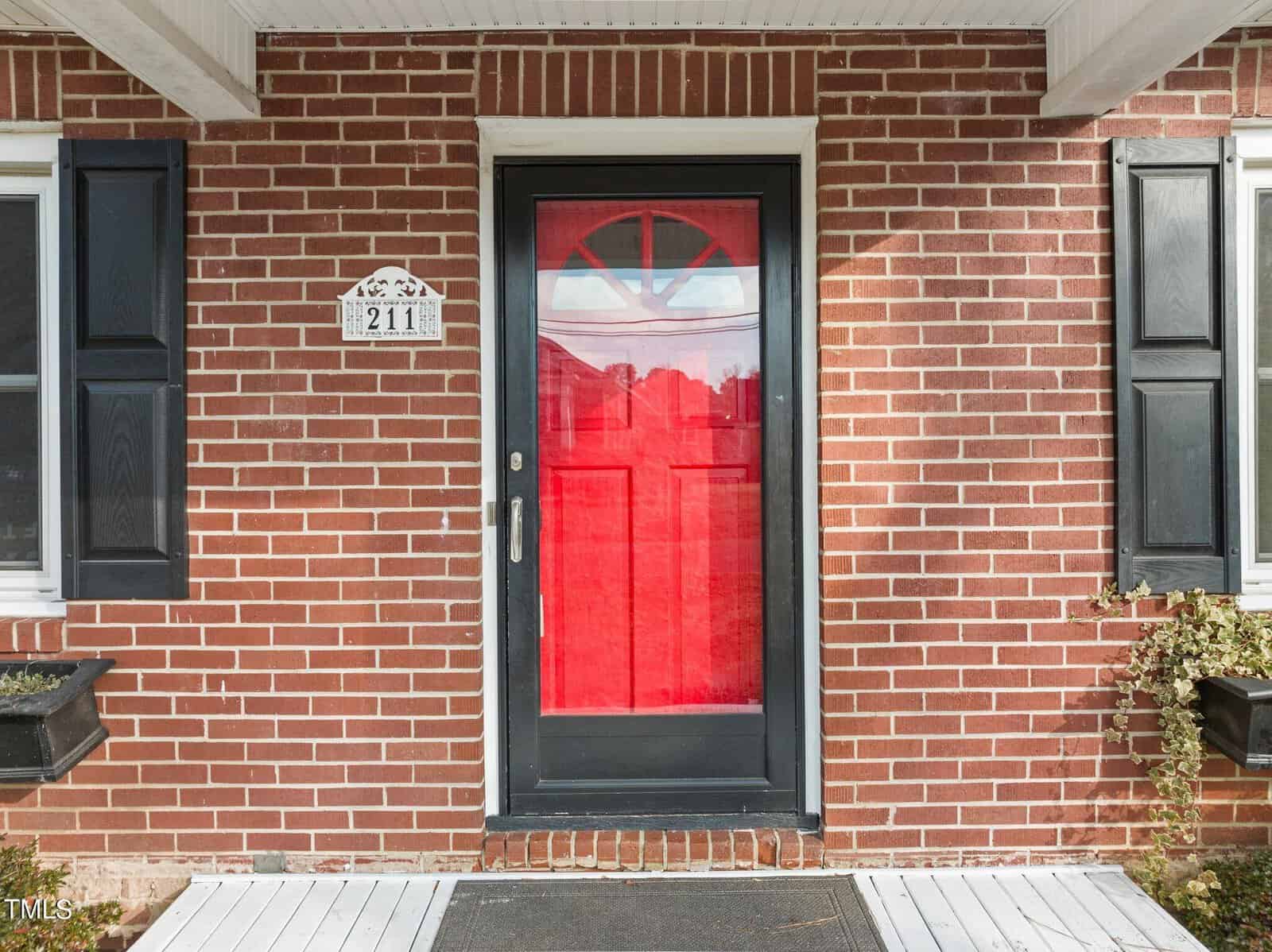 A brick house exterior with a bright red front door framed by black shutters on either side. The house number, 211, is displayed above the door. A small planter with greenery accents the entrance.