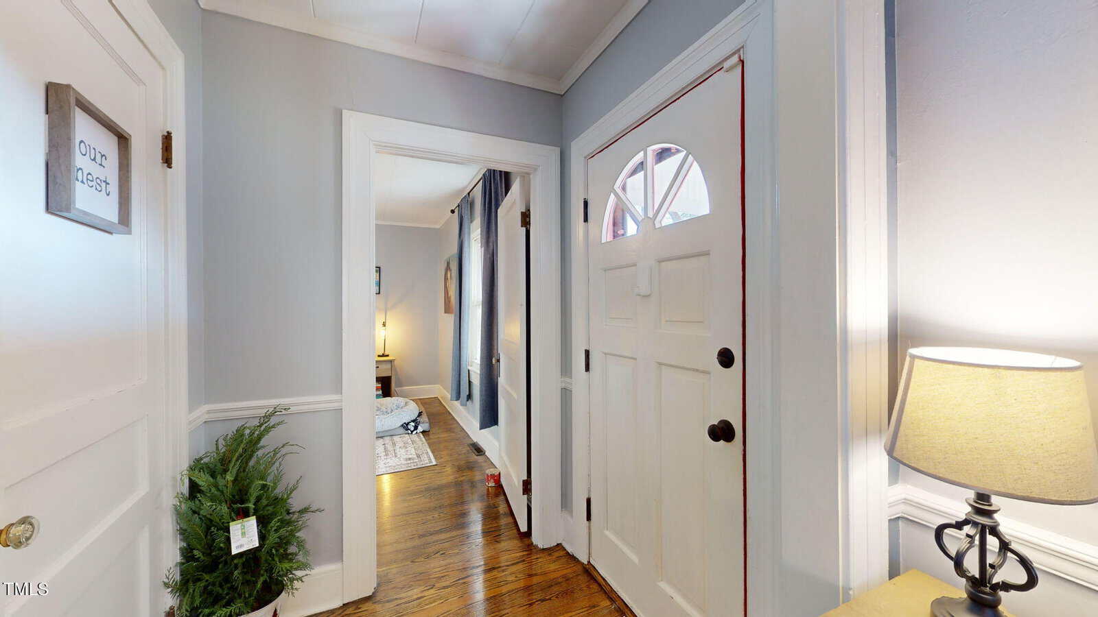 A cozy entryway with a white front door and a small window, illuminated by a nearby lamp on a side table. A potted plant sits on the floor. The hallway leads to another room with wooden floors and light-colored walls.