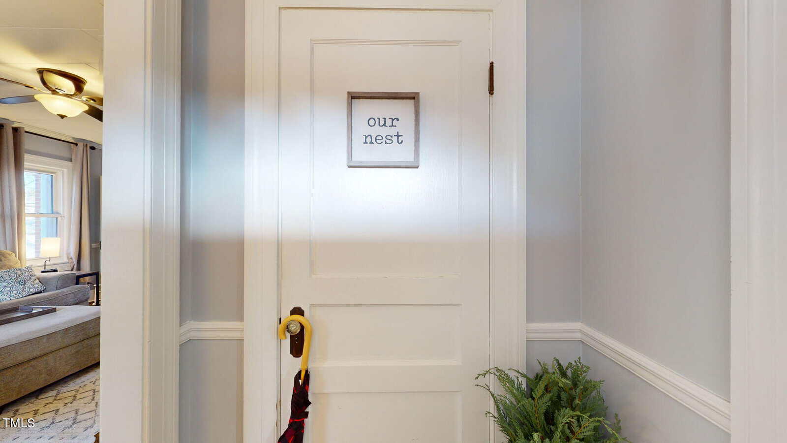 A white door with a framed sign reading "our nest." The door has a handle with a black and red plaid scarf hanging from it. A potted plant is on the floor to the right, and part of a living room is visible through a doorway on the left.