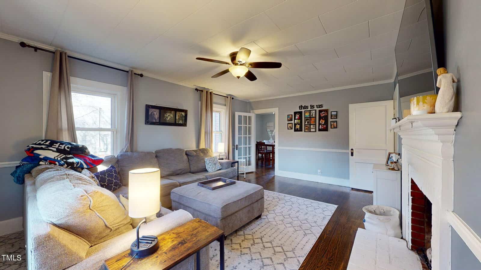 A cozy living room with a gray sectional sofa, a wooden coffee table, and a patterned rug on dark hardwood floors. A ceiling fan hangs overhead. The white mantel displays decor. The walls are gray with family photos and a sign reading "this is us.