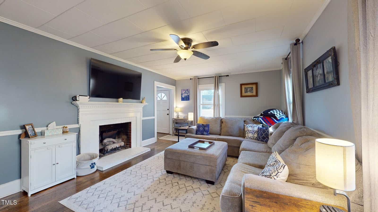 A cozy living room with a gray L-shaped sofa, white brick fireplace, and flat-screen TV. The room features a ceiling fan, wooden floor, and soft lighting with decorative frames on light gray walls and a patterned rug on the floor.