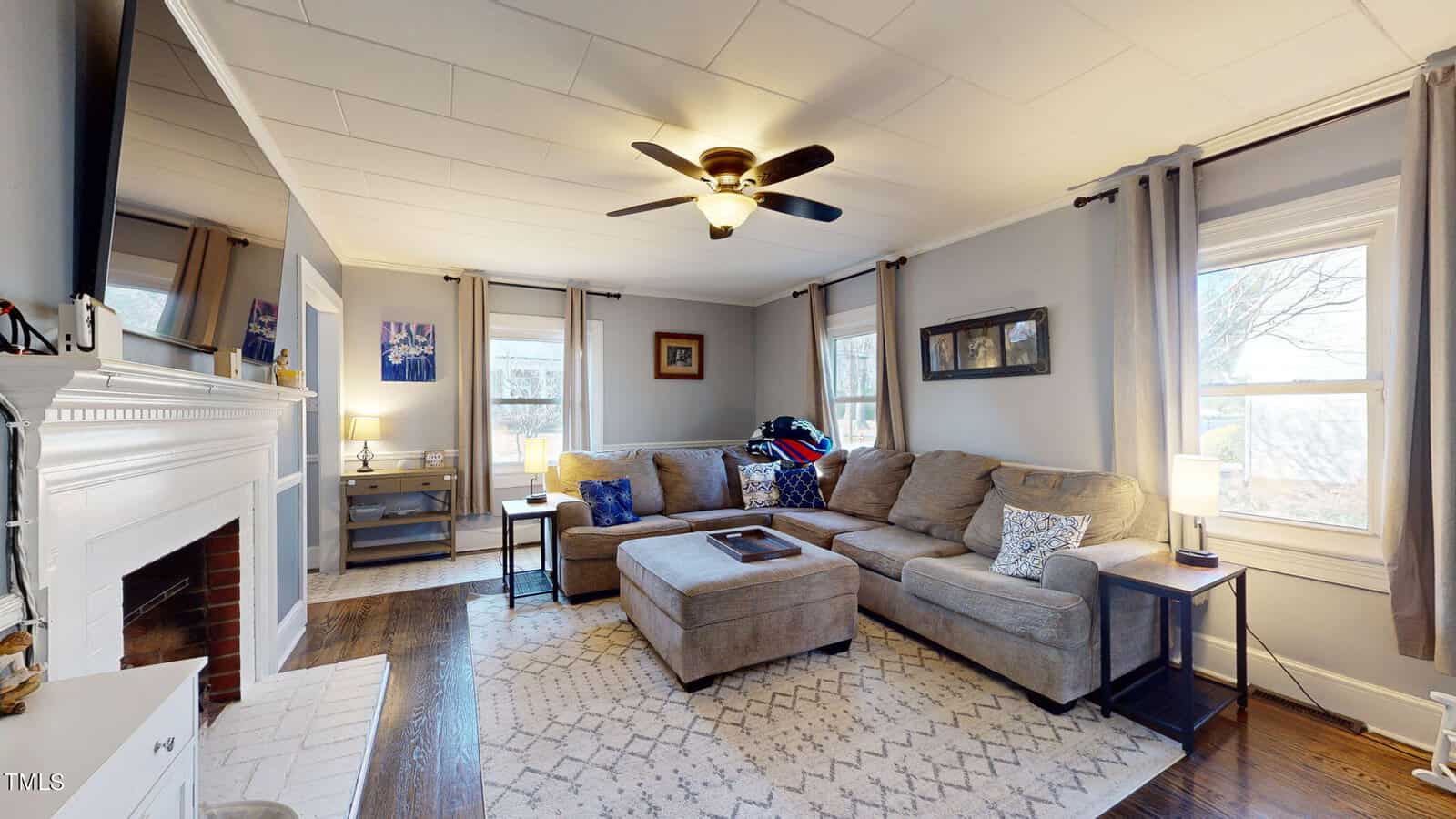A cozy living room with a light brown sectional sofa, patterned cushions, and a white fireplace. The room has a ceiling fan, large windows with curtains, a geometric rug, and framed artwork on the gray walls. A flat-screen TV is above the fireplace.
