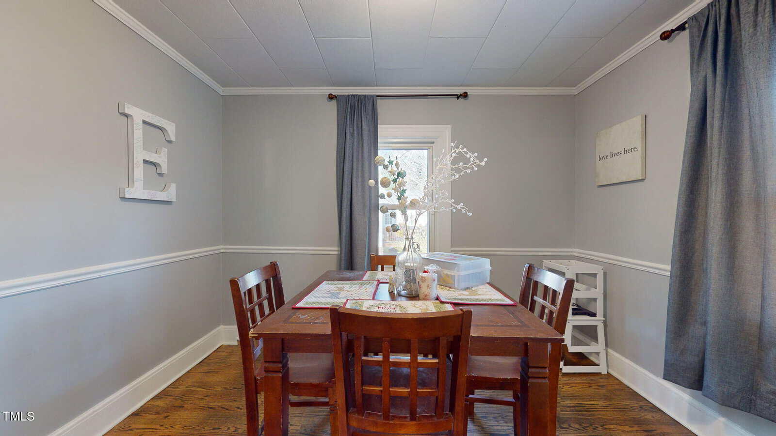 A dining room with a wooden table and four chairs on a wooden floor. The room has gray walls with white trim and a large letter "E" on one wall. A window with dark curtains brightens the space, and a white vase with branches decorates the table.