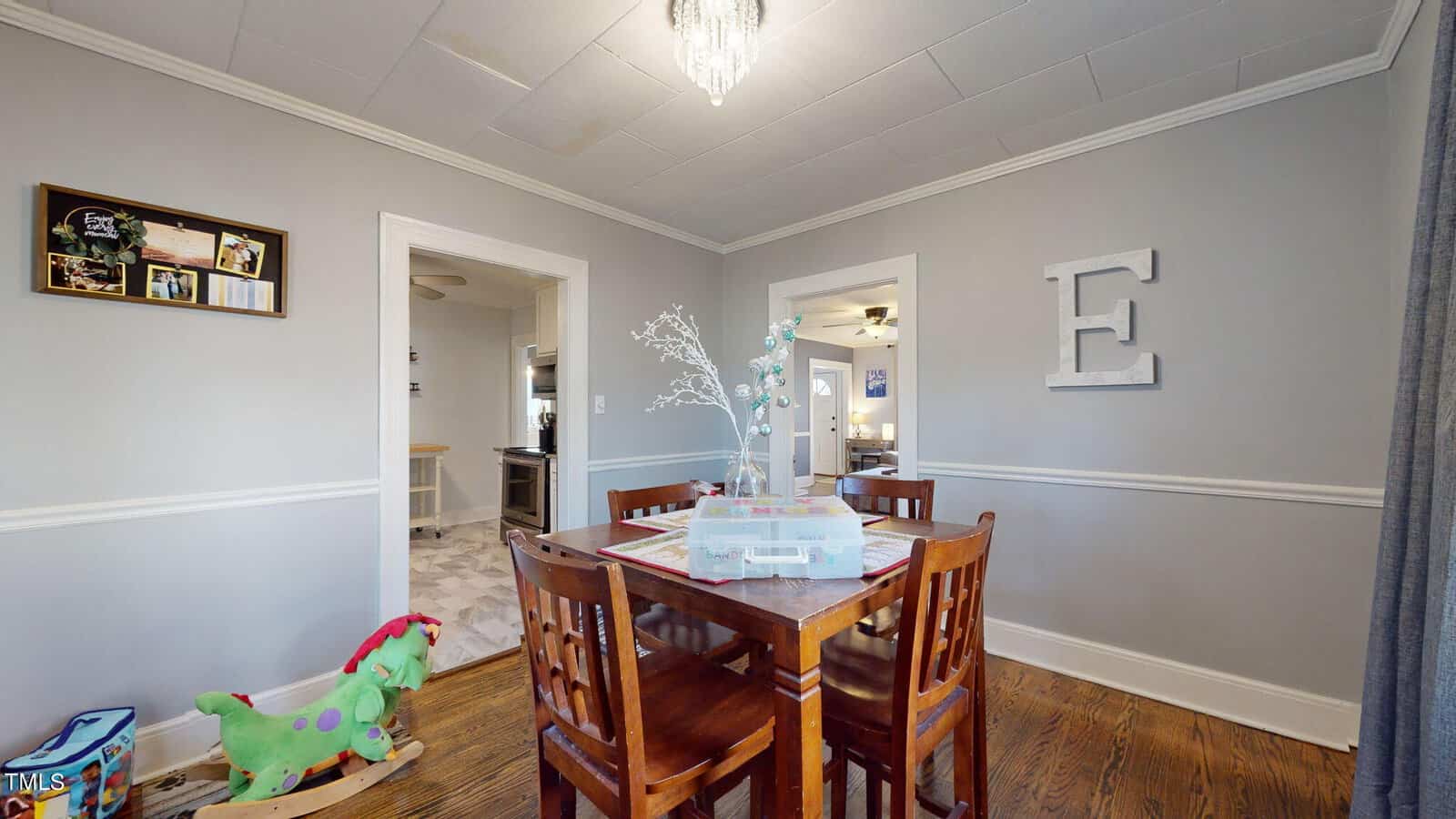 A dining room with a wooden table and four chairs. A small centerpiece is on the table. A colorful rocking horse is on the floor, and a large decorative letter "E" hangs on the wall. A chandelier hangs from the ceiling.