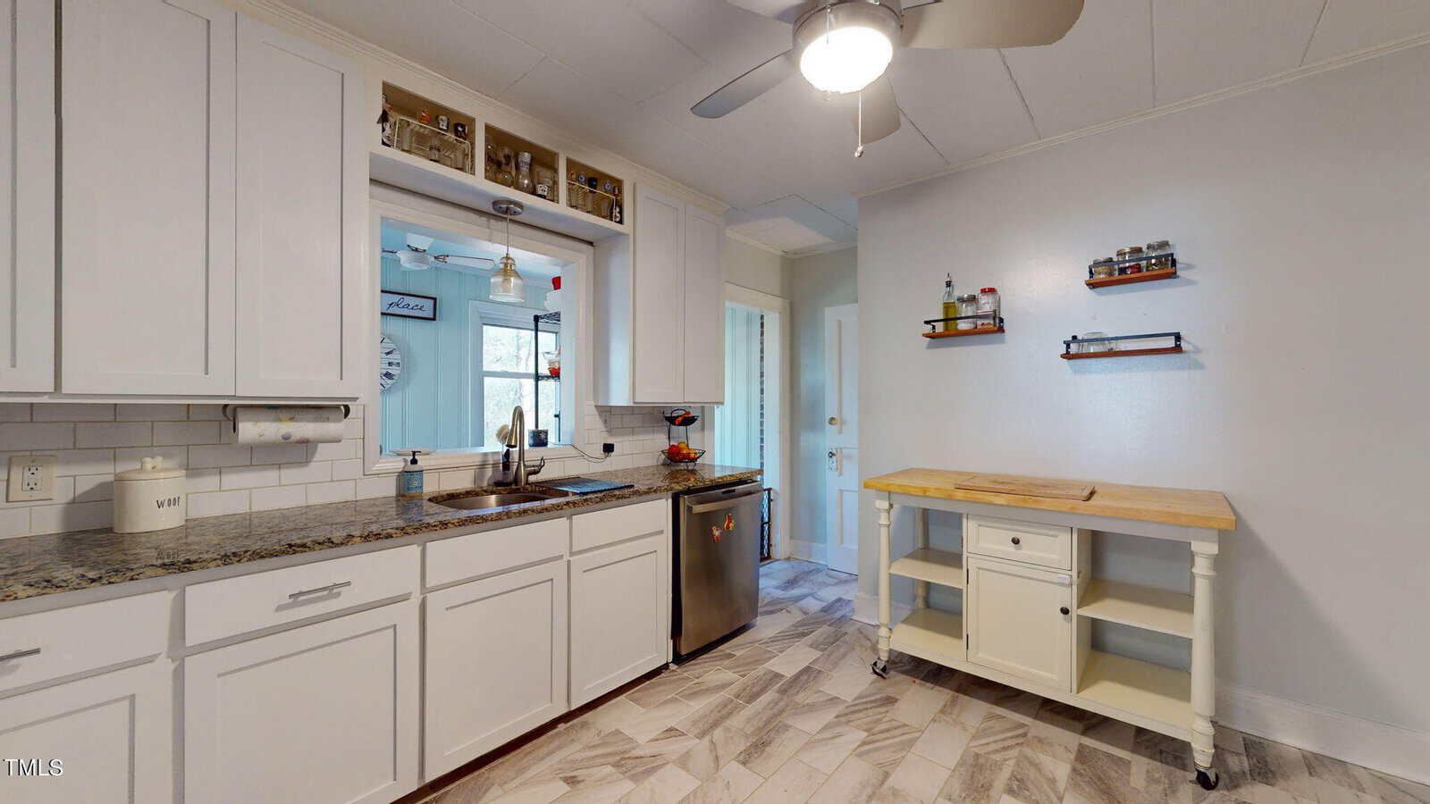 A modern kitchen with white cabinets, granite countertops, and a stainless steel dishwasher. A small kitchen island with shelves and a cutting board is on the right. Overhead, a ceiling fan with a light illuminates the room.