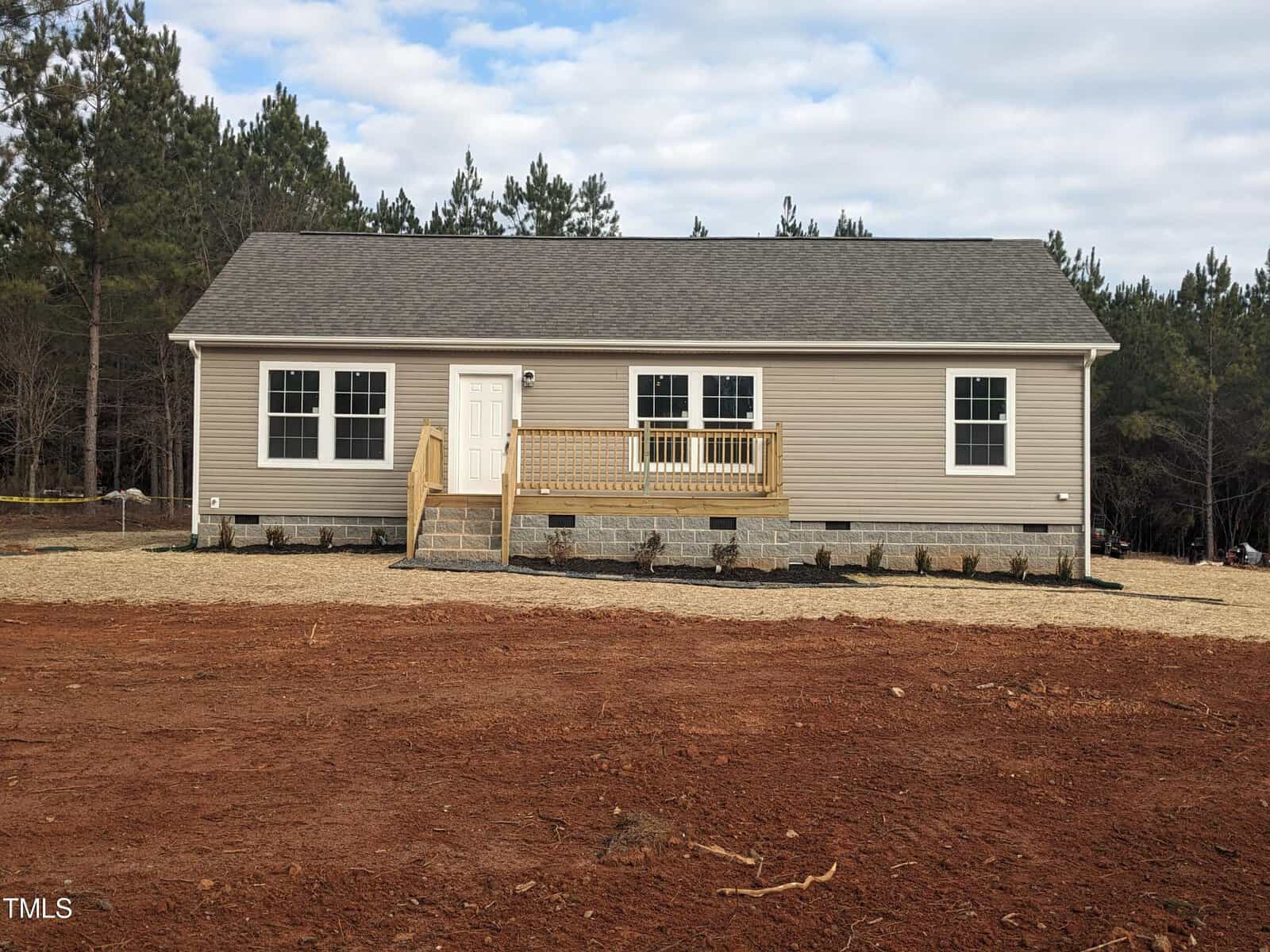 A new single-story home with beige siding, white trim, and a small front deck, situated on a cleared lot with red soil and sparse greenery.