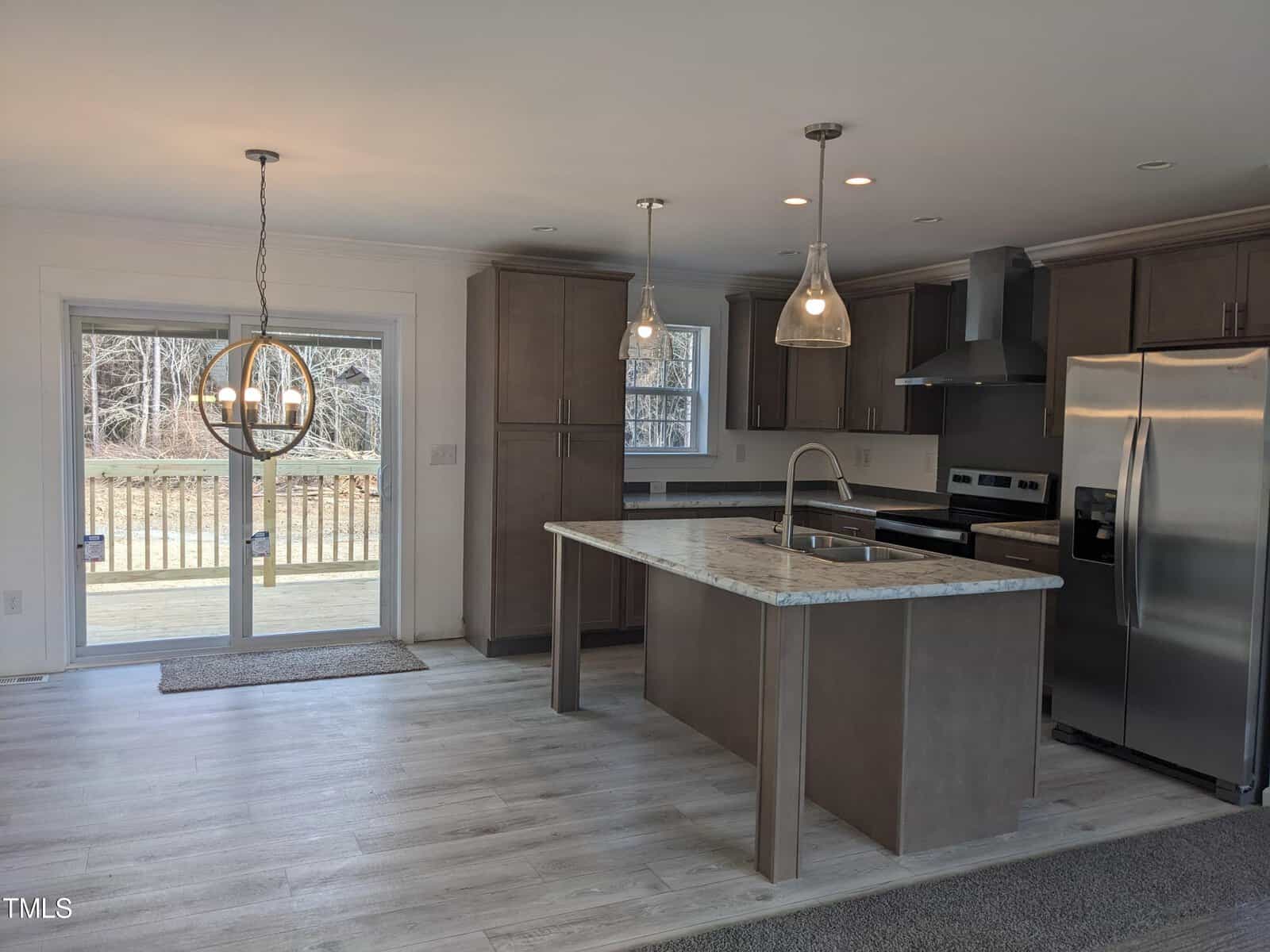 Modern kitchen with gray cabinetry and stainless steel appliances. features an island with a marble countertop, two pendant lights, and a view of the backyard through large glass doors.