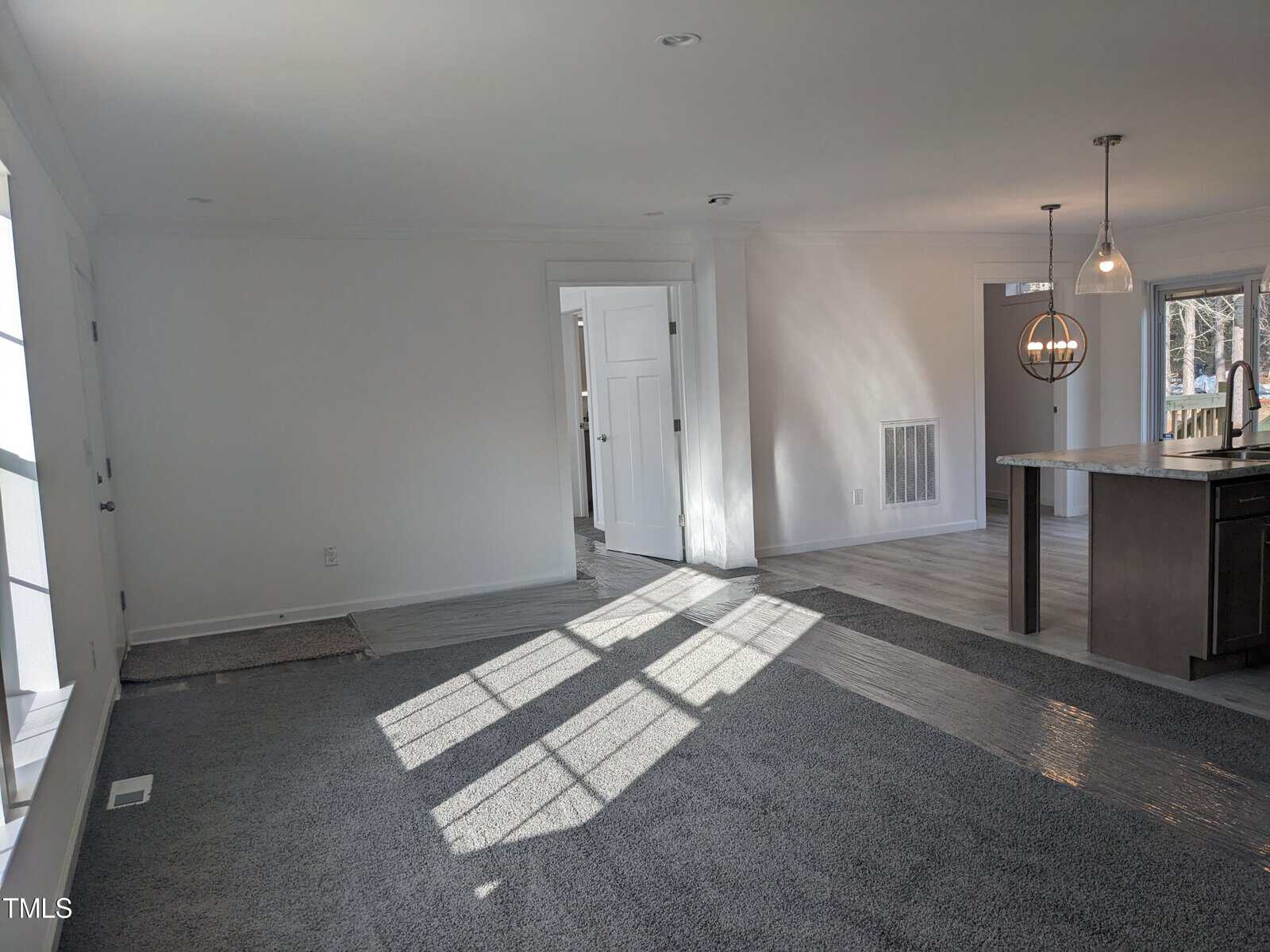 Interior of a modern home featuring a room with gray carpet and walls, sunlight casting shadows on the floor, an open doorway, and a glimpse of a kitchen with a person standing.
