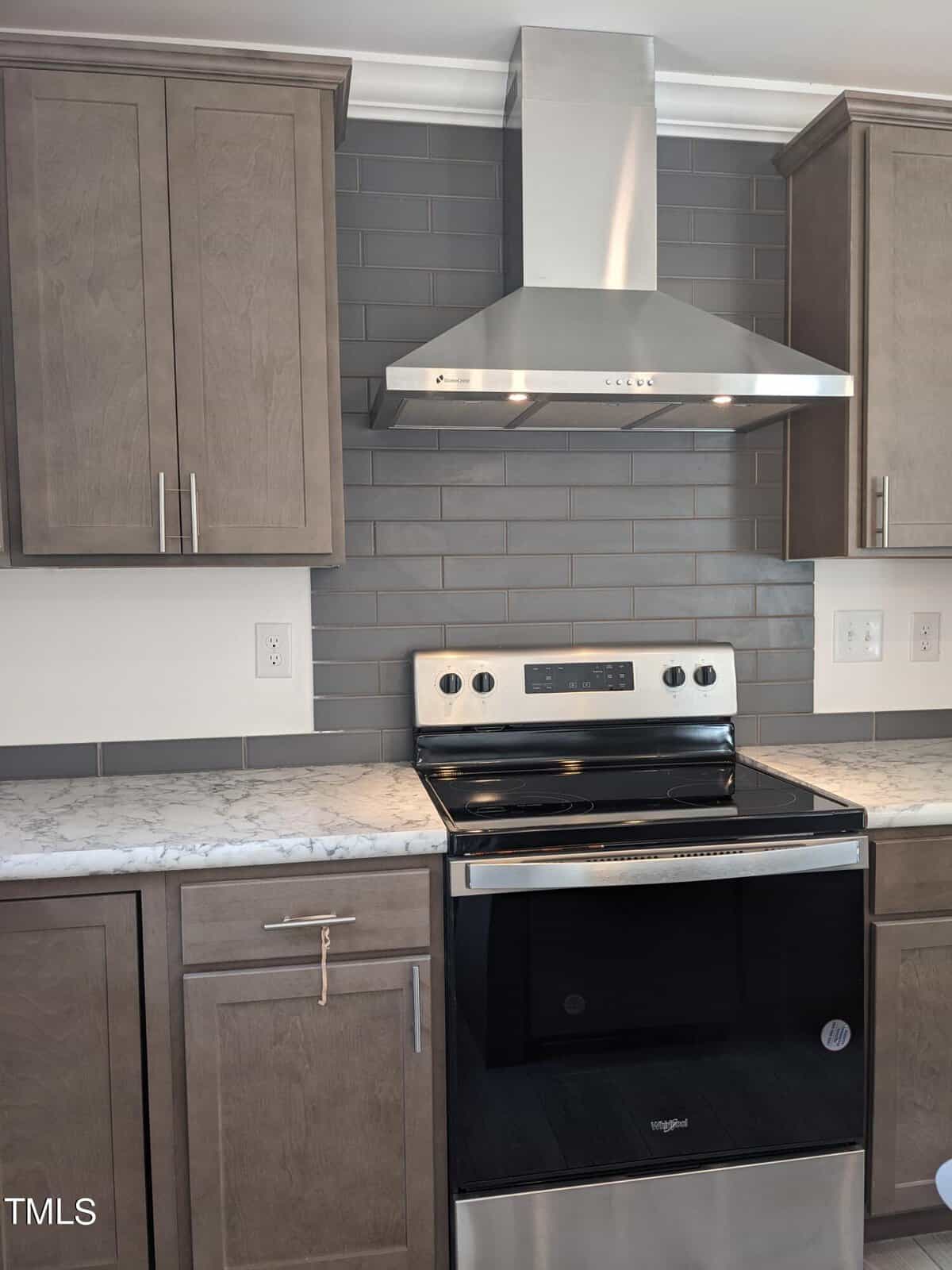 A modern kitchen featuring gray cabinets, a stainless steel range with an oven, and a matching overhead exhaust hood. the backsplash is tiled in dark gray and the countertops are light gray marble.