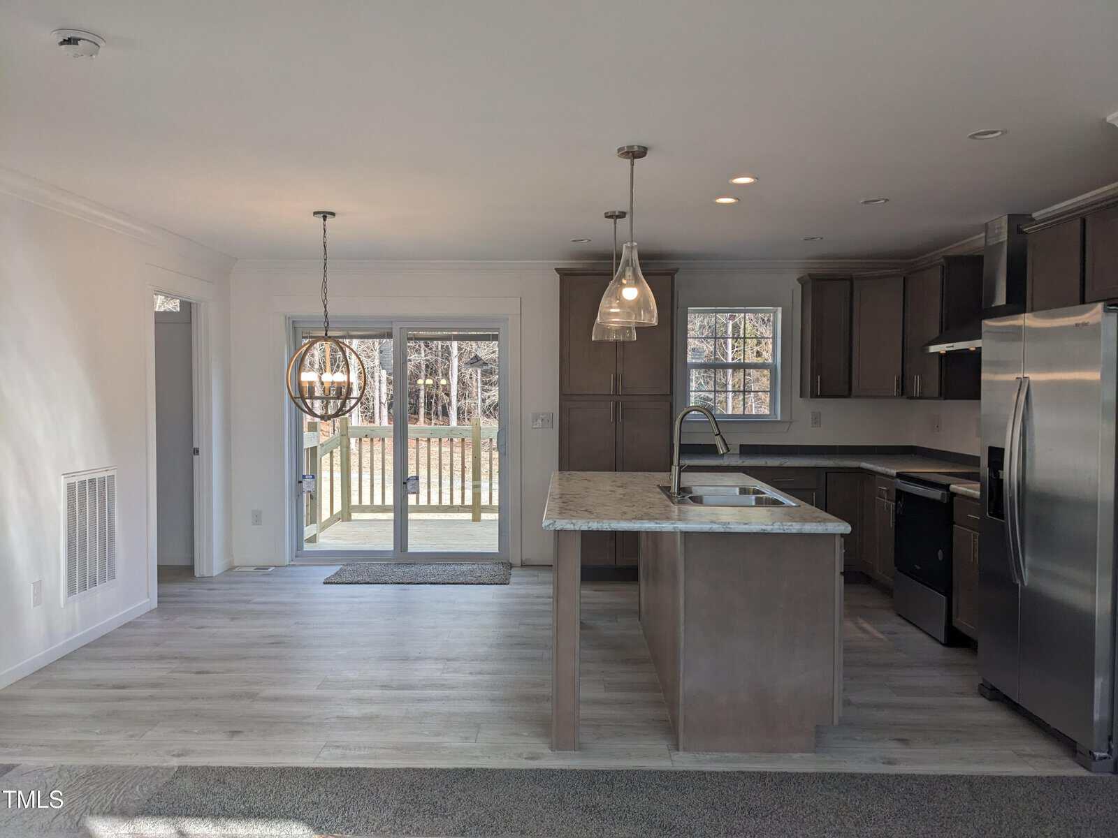 A modern kitchen with dark wood cabinets, stainless steel appliances, and a center island with a sink, leading to a bright dining area with large windows and a door to the backyard.