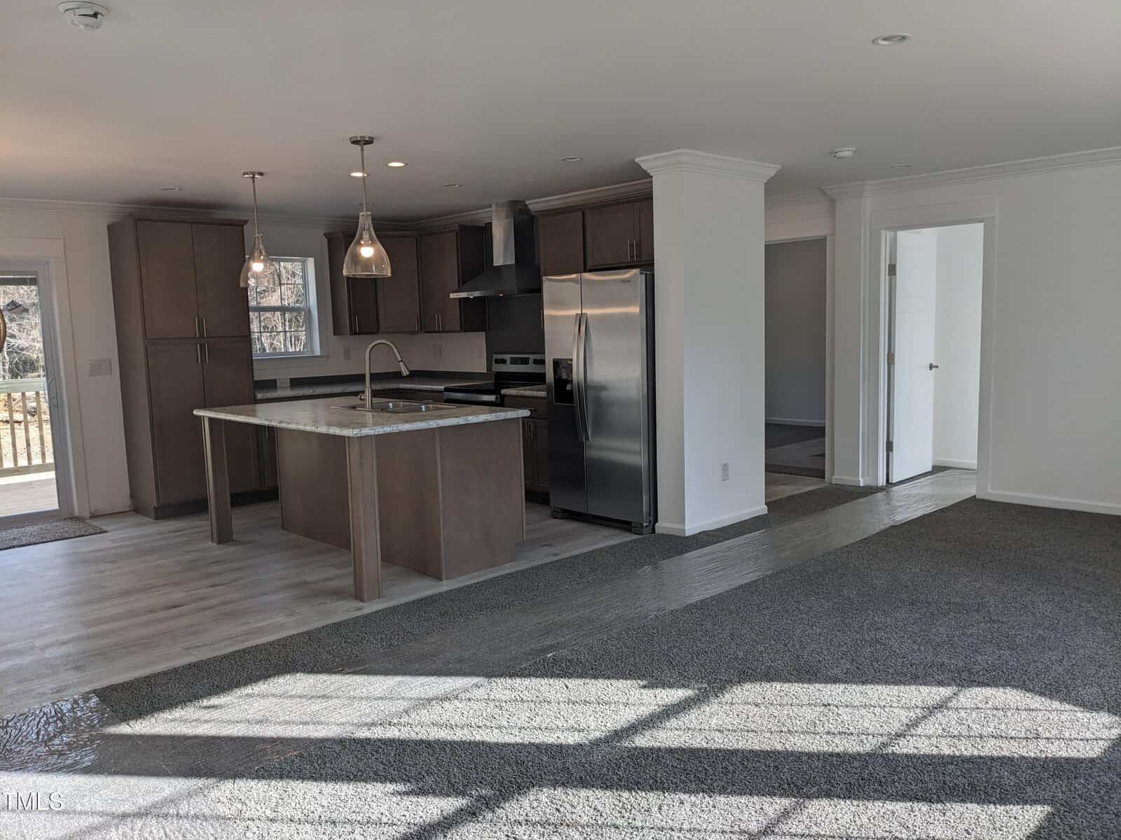 A modern kitchen with wooden cabinets, stainless steel appliances, and a center island under natural light, showcasing a mix of hardwood and tiled flooring.