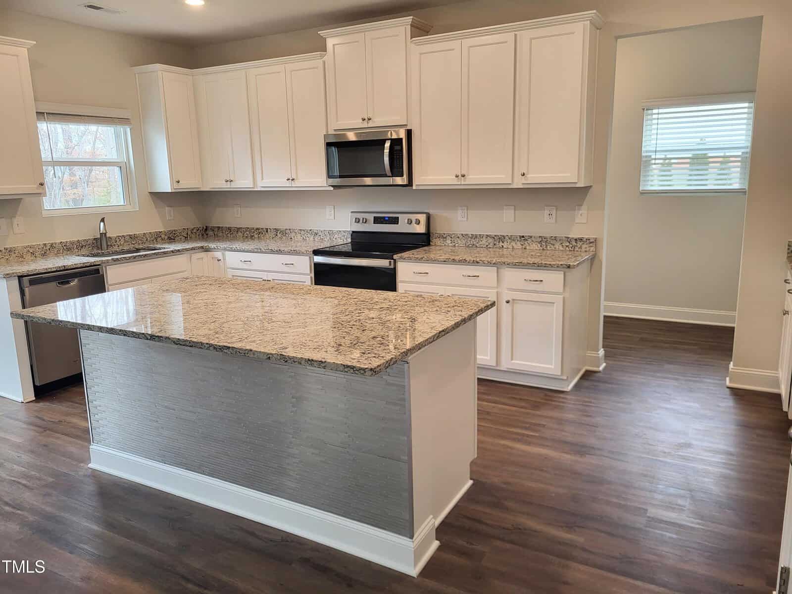 A modern kitchen with white cabinets, granite countertops, and stainless steel appliances. There's a large center island with a textured front panel. The floor is dark wood, and the room is well-lit with natural light from a window.