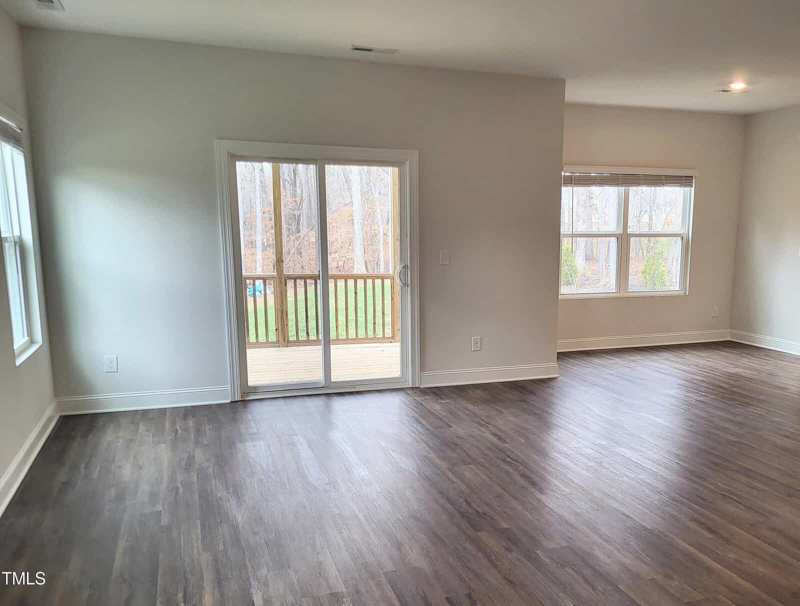 An empty room with wooden flooring and light-colored walls features a sliding glass door leading to a wooden deck. Two large windows provide a view of the trees outside, allowing natural light to fill the space.