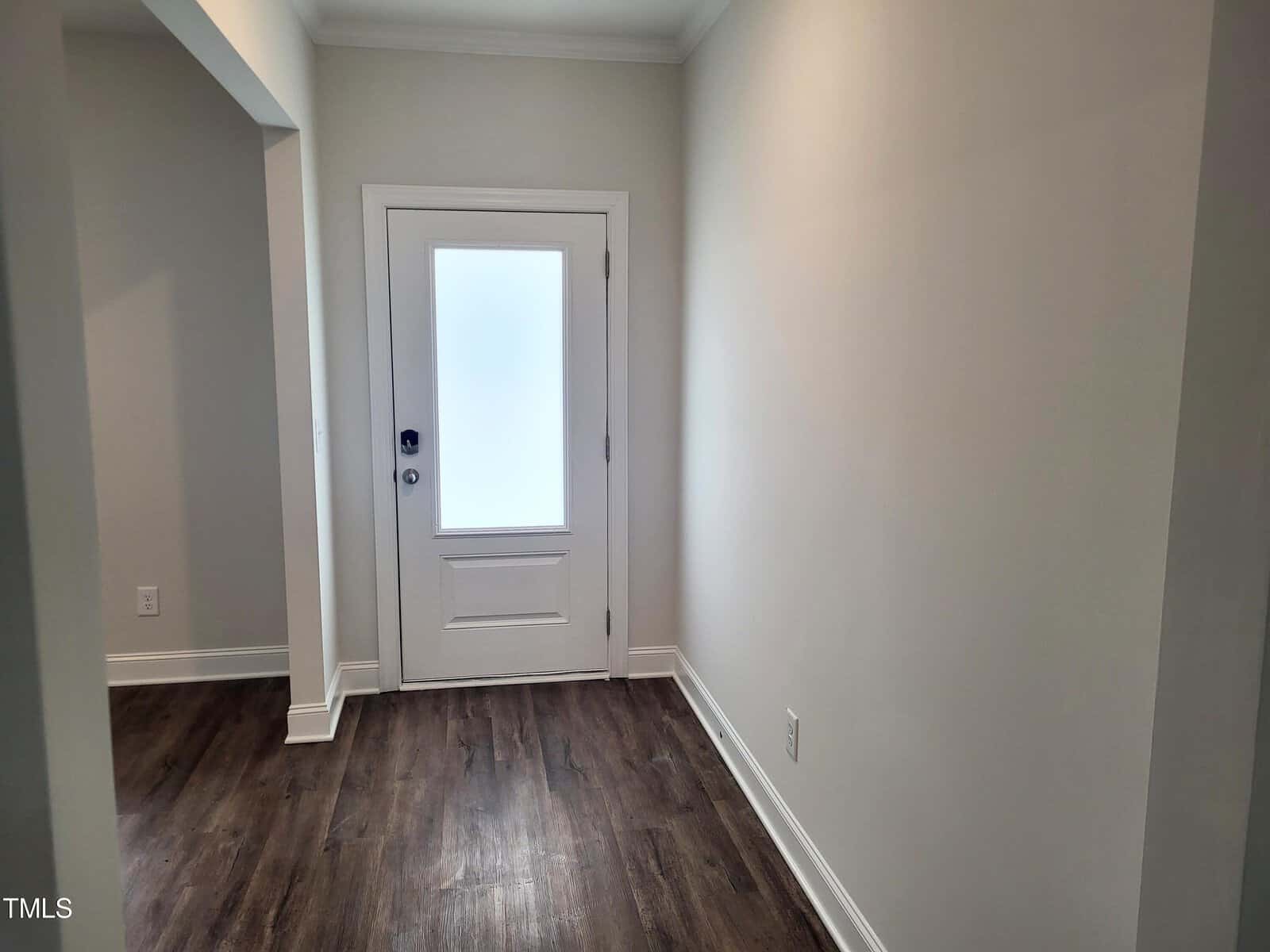 A narrow, well-lit hallway with light-colored walls and dark wood flooring. At the end of the hallway is a white door with a frosted glass panel, allowing diffused light to enter. The hallway leads to an open doorway on the left.