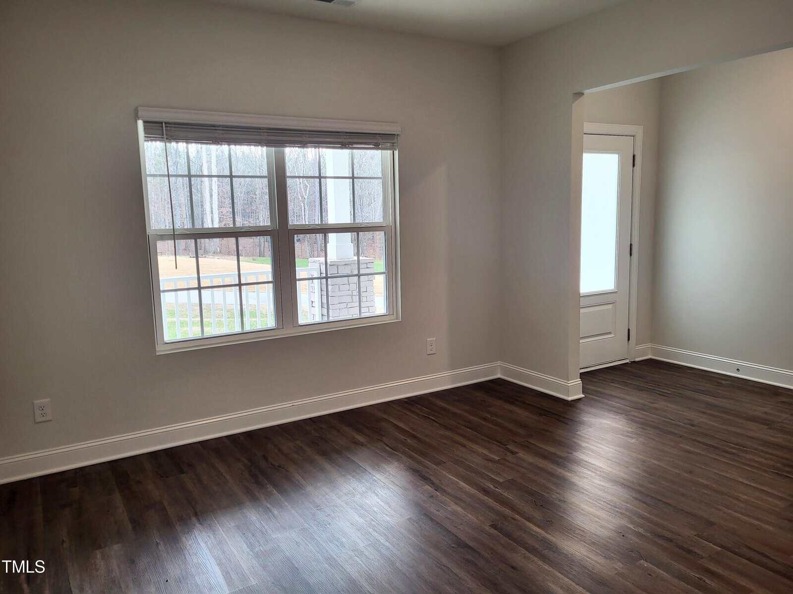 Bright, empty room with large window, brown wooden floor, and light-colored walls. The window overlooks a yard, and there's a partially open door on the right side leading to another room.