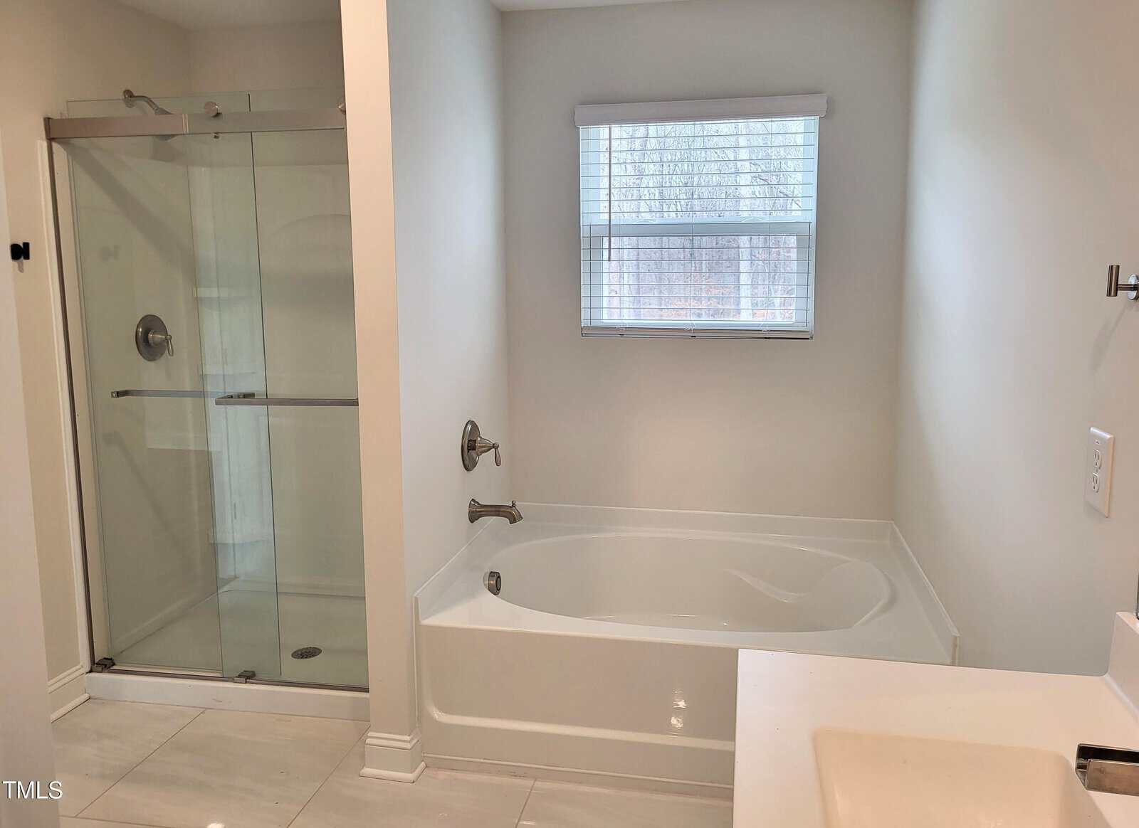 A bathroom with a white bathtub, glass-enclosed shower, and a window with blinds. The walls and floor are in light colors, providing a clean and modern look.
