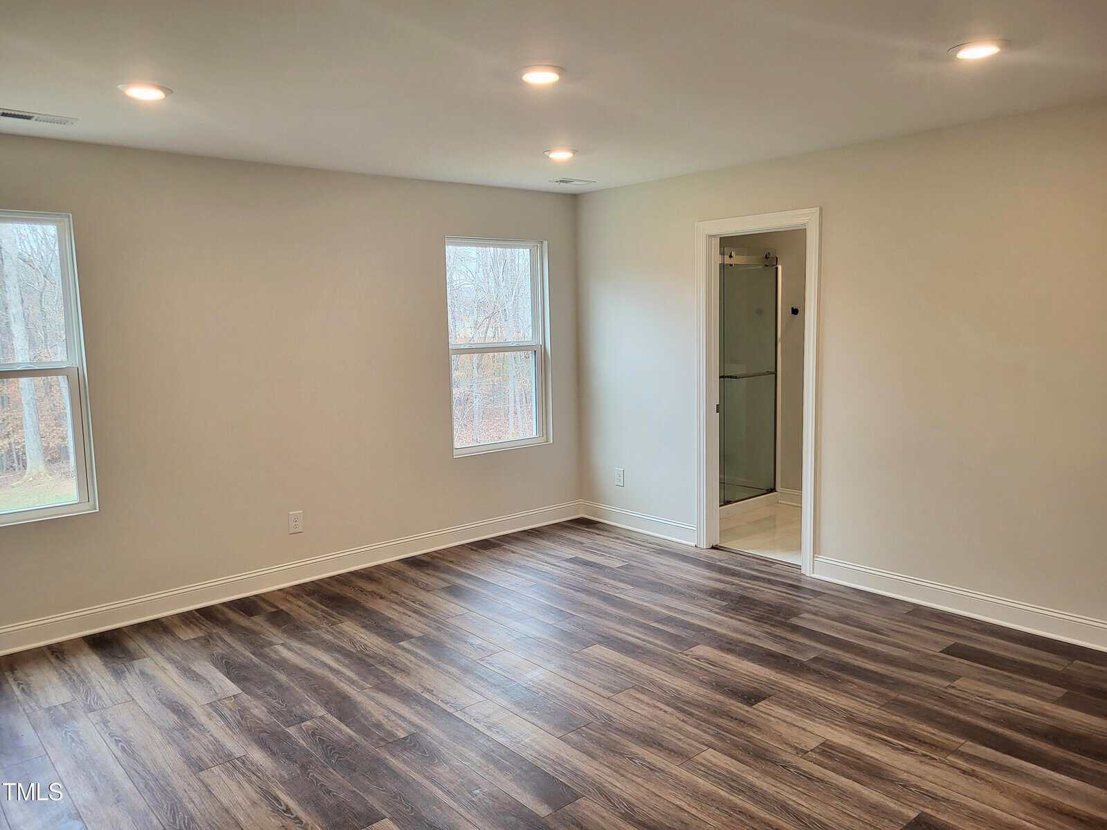 A bright, empty room with light gray walls and wood-look flooring. Two windows let in natural light. An open door on the right leads to a bathroom with a glass shower. Recessed ceiling lights illuminate the space.