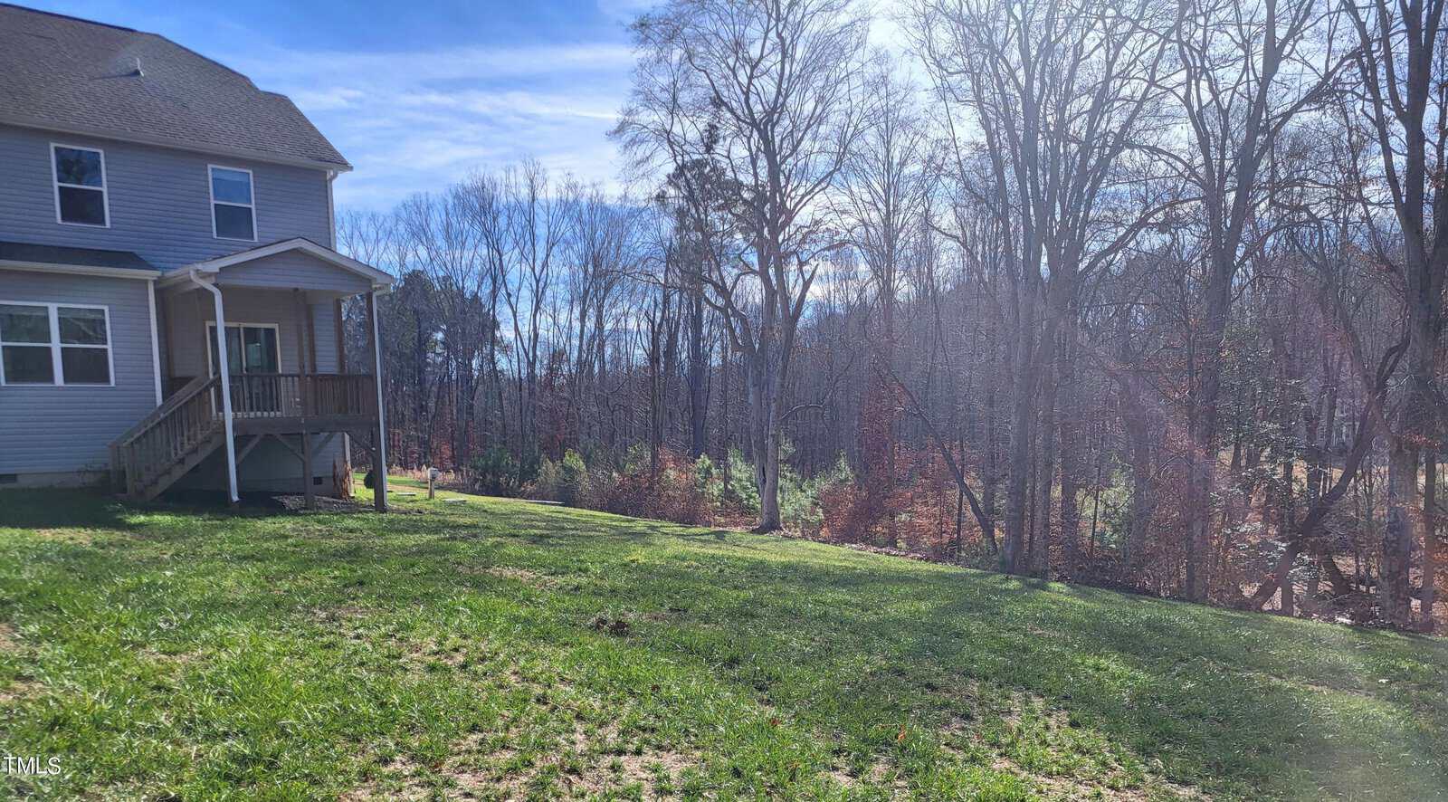A two-story house with a small porch sits alongside a grassy lawn. The background features a forest of leafless trees under a clear blue sky. Sunlight streams through the trees, creating a serene and peaceful atmosphere.