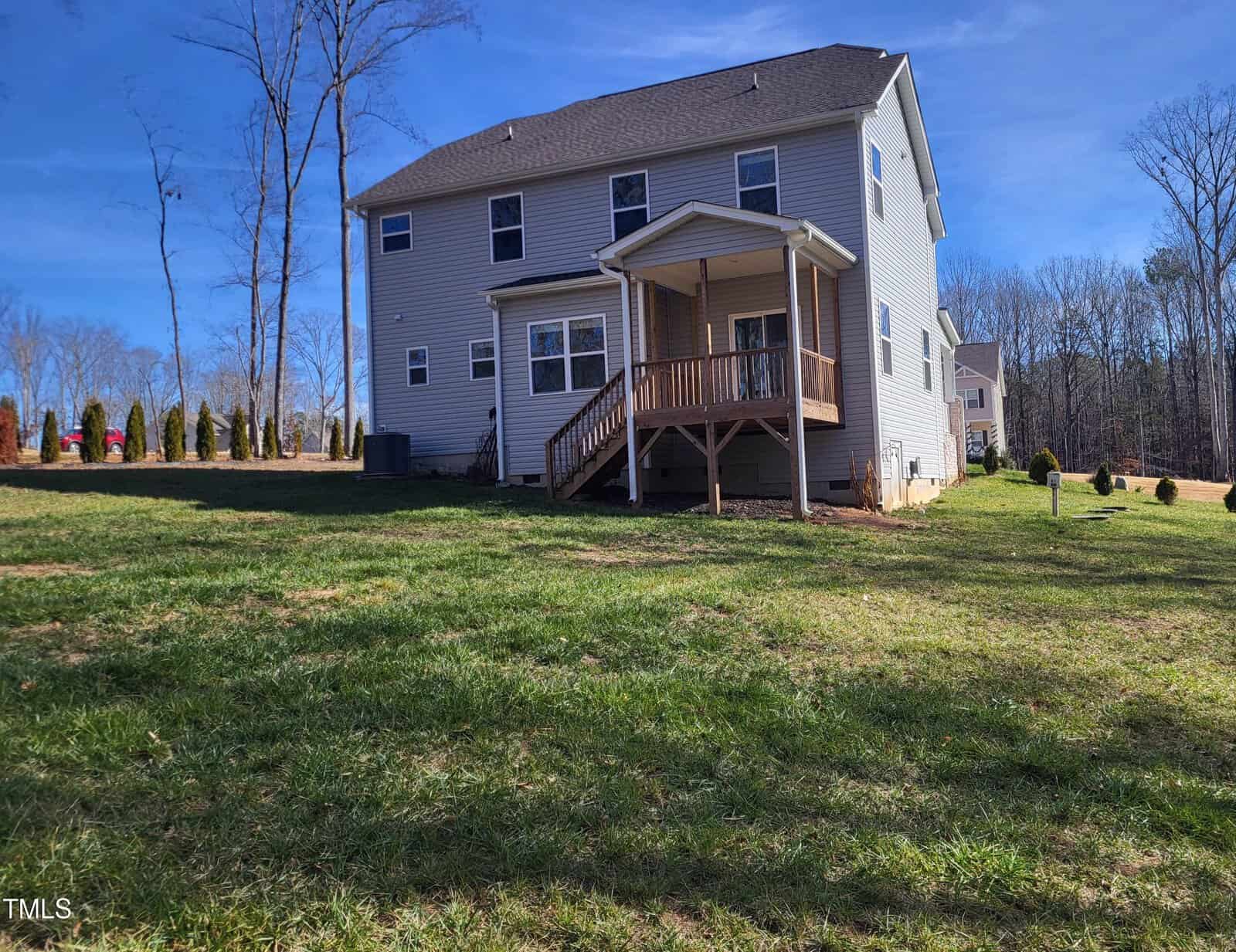 A two-story house with a beige exterior and a wooden porch is situated on a grassy lawn. Bare trees surround the area under a clear blue sky.