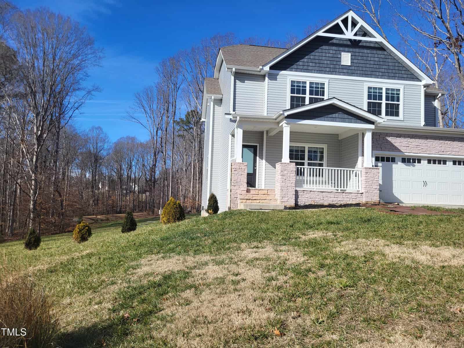 A two-story gray house with white trim at 3447 Lilac Lane features a covered porch and a garage, nestled on a grassy slope in Wake Forest. Small shrubs and trees surround the home, while the clear blue sky indicates a sunny day.