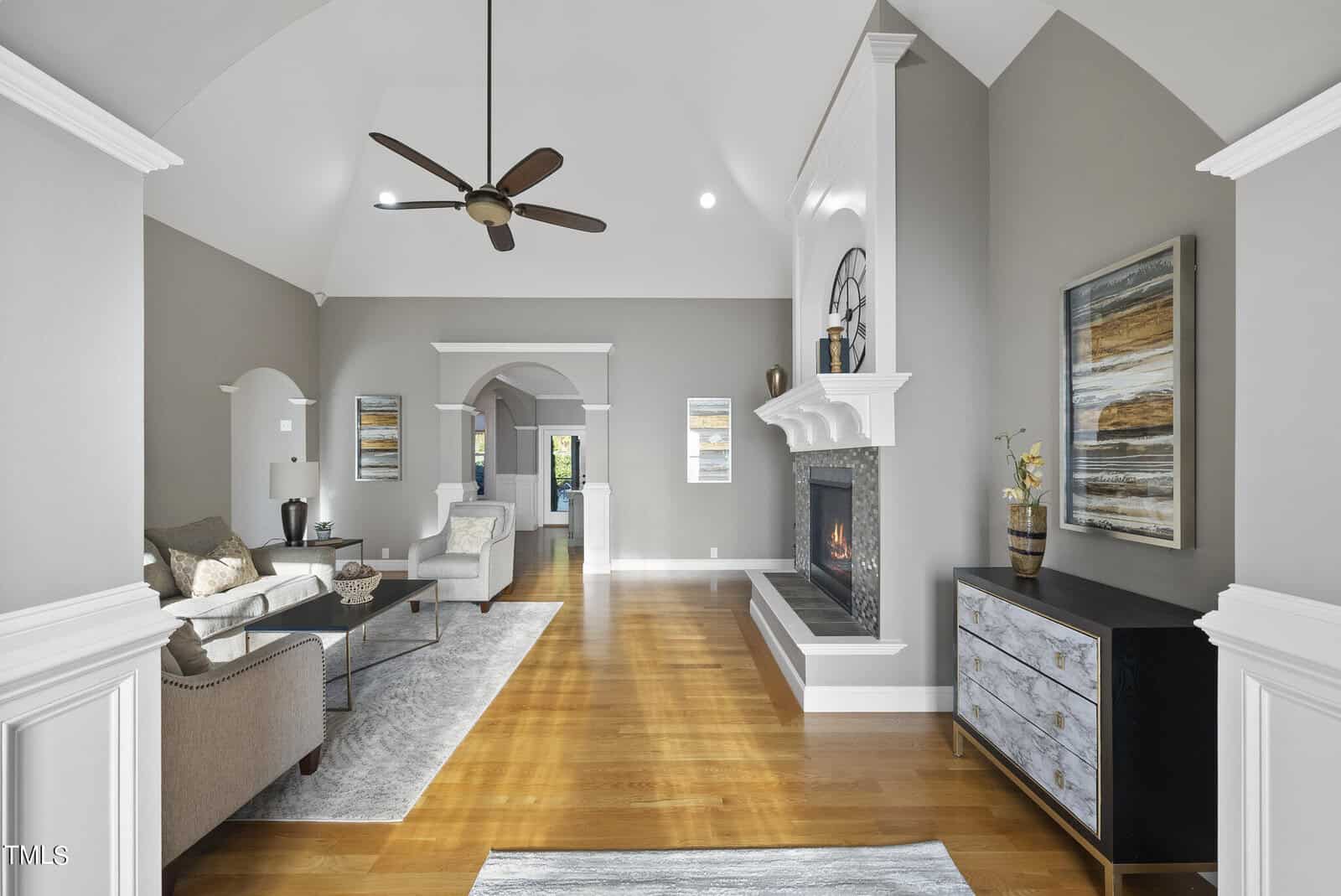 Nestled at 1107 Dovefield Lane in Youngsville, this spacious modern living room boasts high ceilings and hardwood floors. A cozy fireplace, neutral-toned furniture, abstract wall art, and a ceiling fan complement the space as light pours in from a distant window, enhancing the elegant atmosphere.