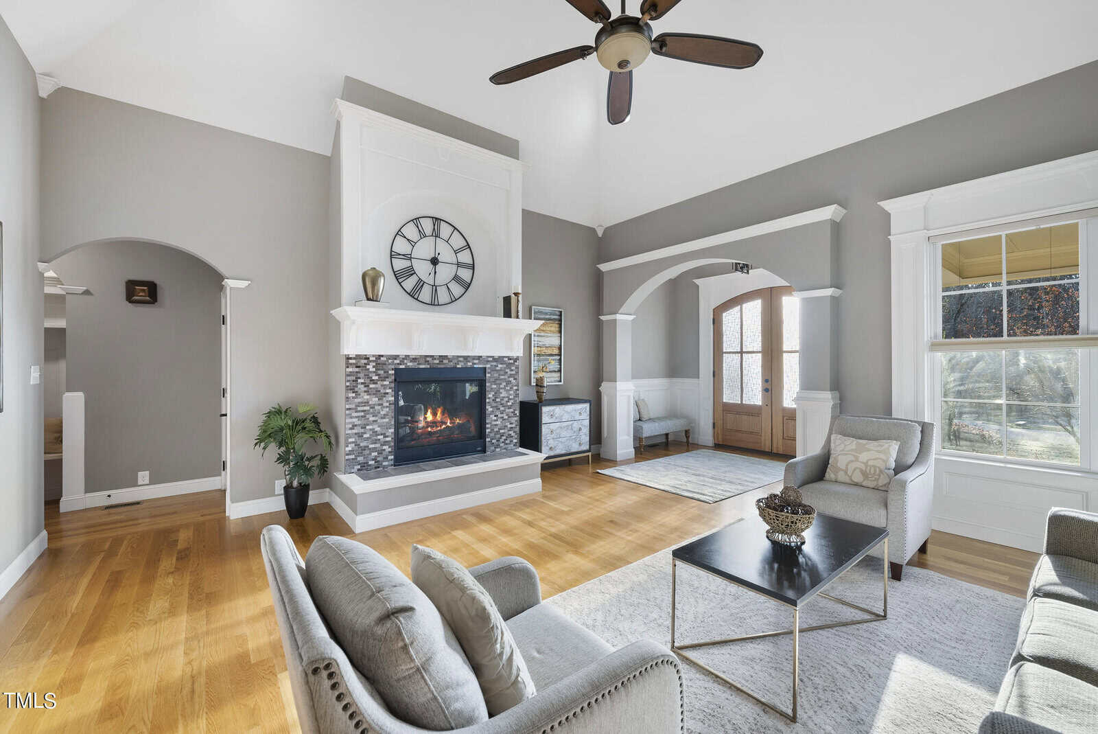 A spacious living room with a vaulted ceiling features a large clock above a modern fireplace. Two armchairs and a sofa surround a coffee table, with light wood flooring and arched doorways adding elegance. A ceiling fan is overhead.