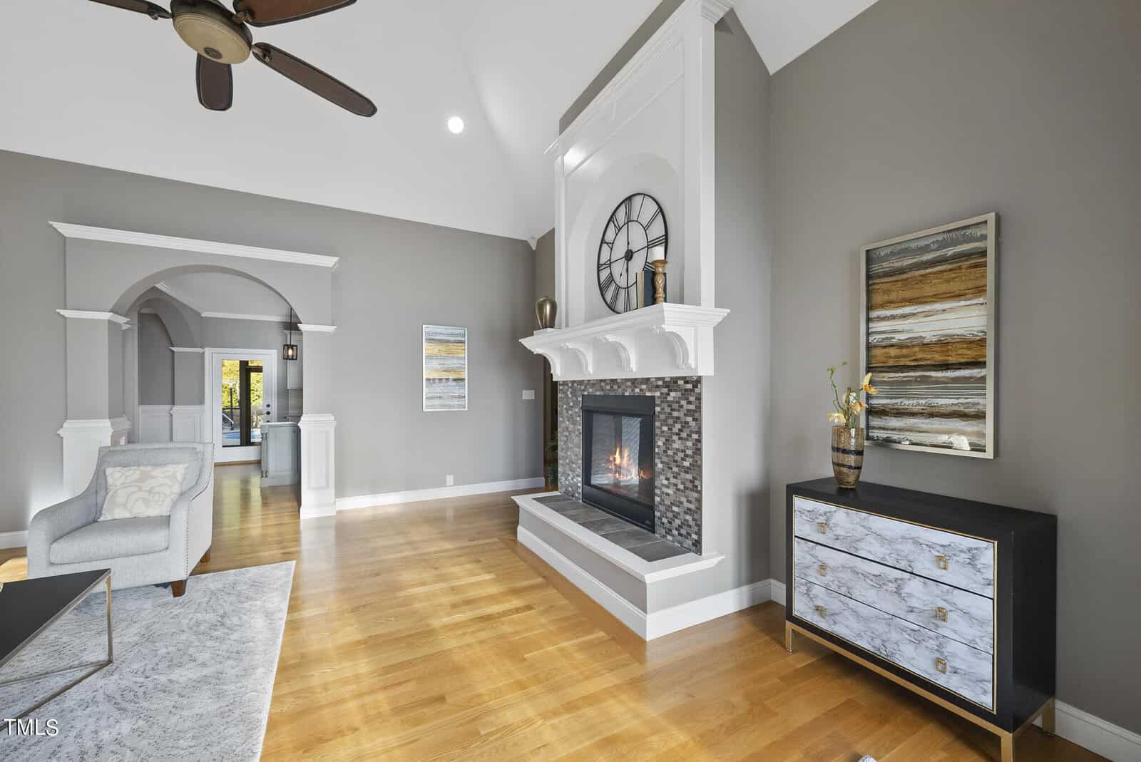 A modern living room with hardwood floors, a tiled fireplace, and a large clock above the mantel. It features a light gray armchair, a colorful abstract painting, and a dark dresser with decorative items. An arched doorway leads to another room.