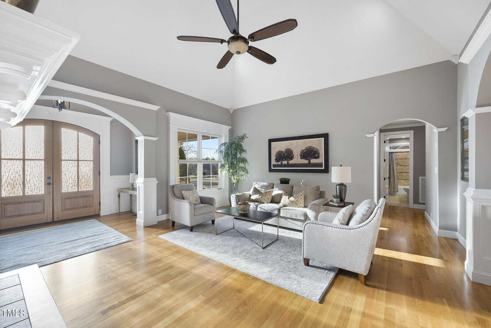 Spacious living room with high ceiling, large fan, and light wood flooring. Gray walls feature a picture and arched doorway leading to a hallway. Two sofas and a glass coffee table center the space, with natural light streaming through large windows.