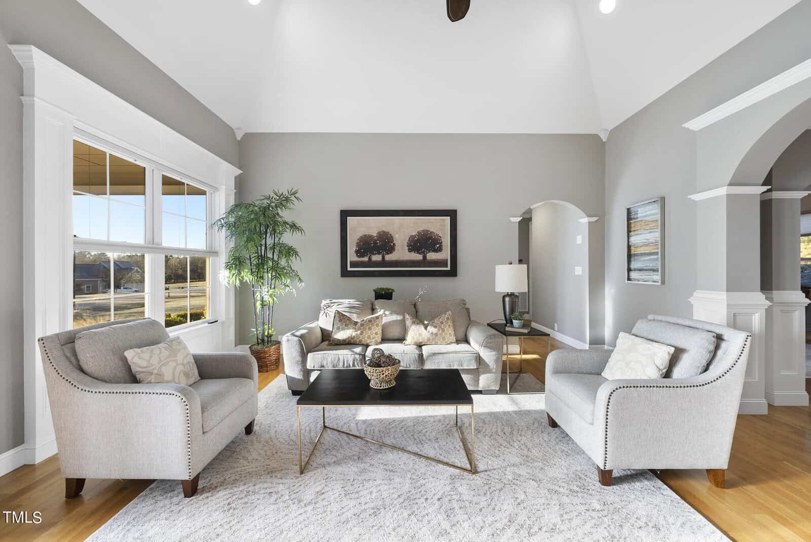A bright, elegant living room with high ceilings. It features a beige sofa, two gray armchairs, a modern coffee table, and a large window with views. The room has a neutral color palette and a framed nature painting on the wall.