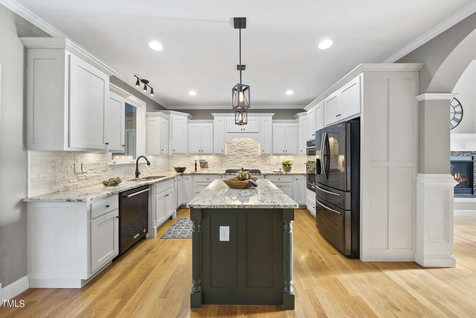 A modern kitchen with a central island featuring a granite countertop and dark base. It has white cabinets, stainless steel appliances, a double sink, and wooden flooring. Pendant lighting illuminates the space, complementing recessed ceiling lights.