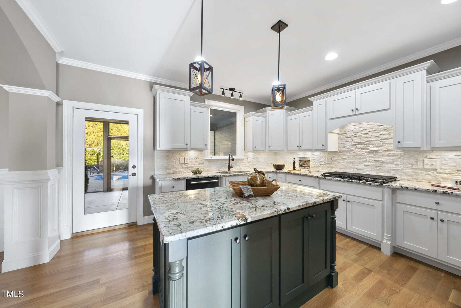 A modern kitchen with white cabinets, a large granite island with a dark base, and pendant lights overhead. Hardwood floors and a glass door leading to an outdoor area are visible.