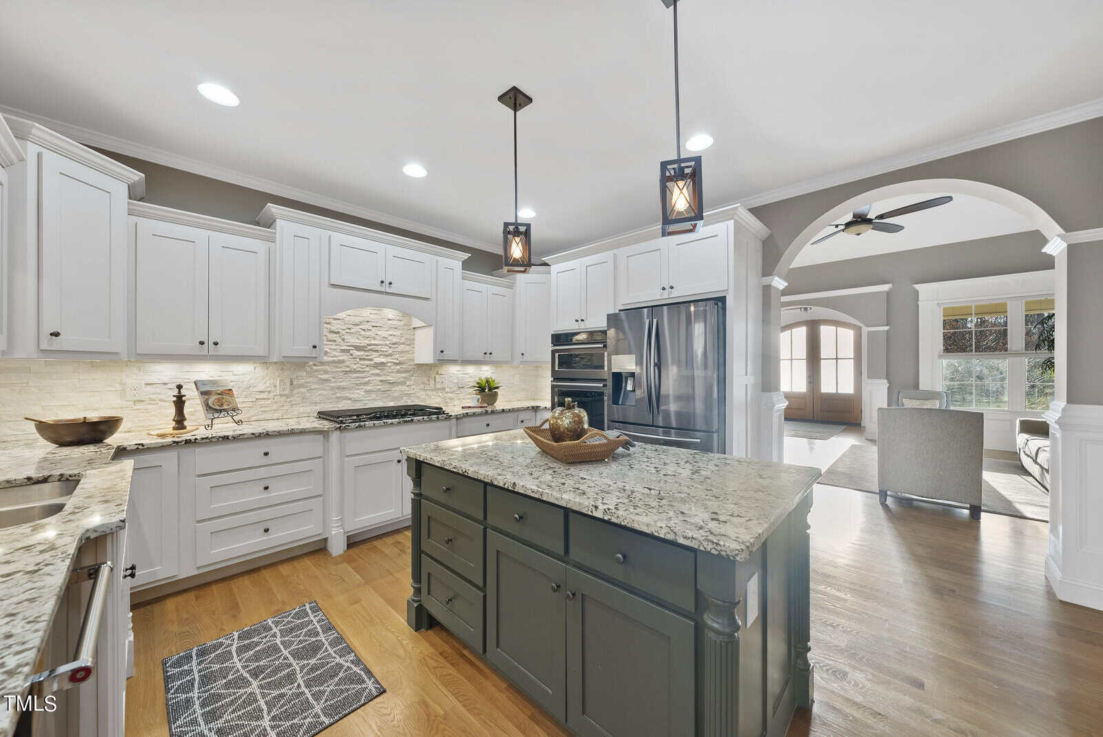 Spacious kitchen with white cabinets, granite countertops, and a large dark green island. Modern appliances and pendant lighting are visible. An open archway leads to a living area with a ceiling fan and sunlit windows. Hardwood floors throughout.