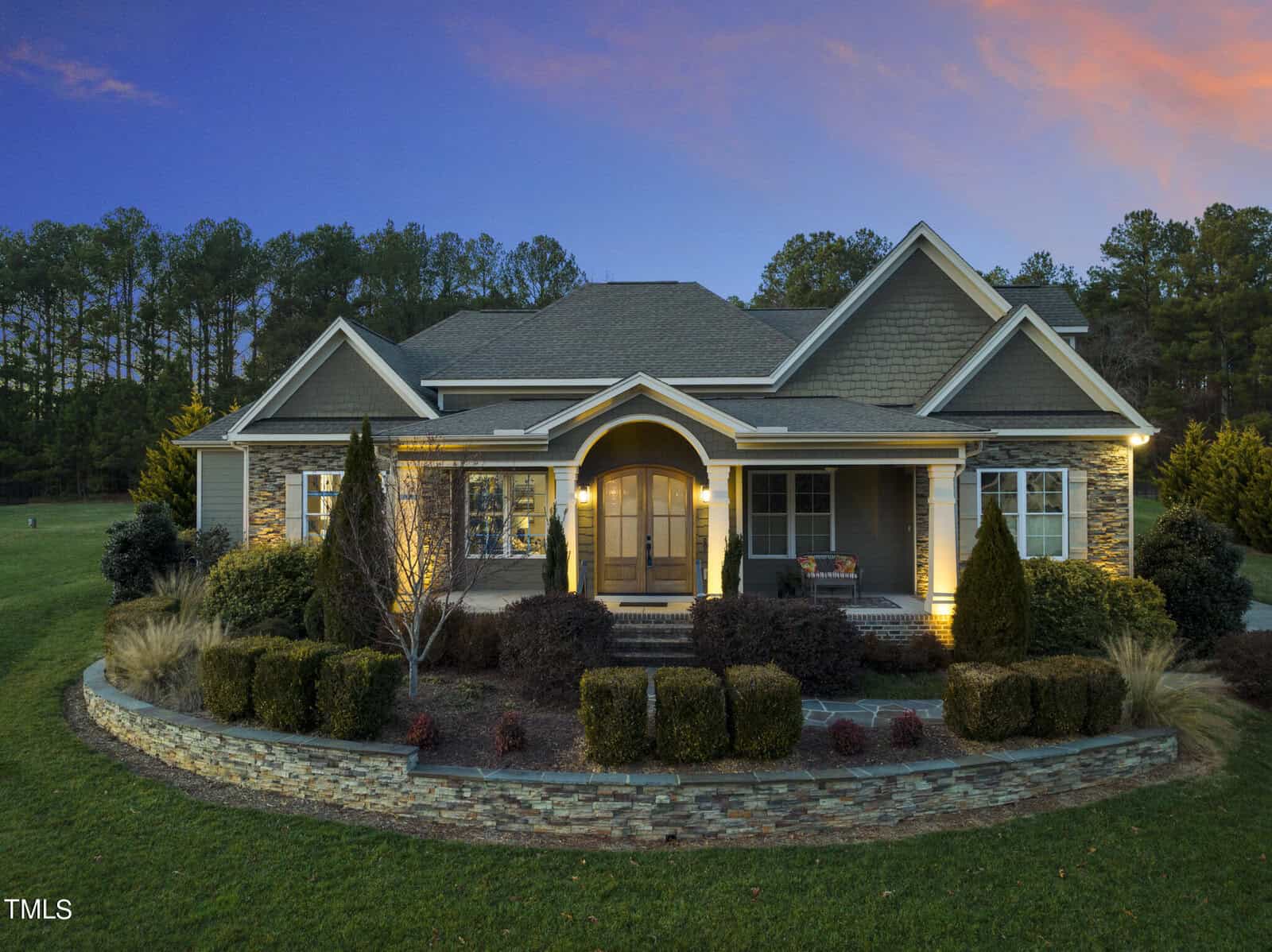 A single-story house with a stone facade and warm exterior lighting at dusk. It features a covered porch, well-maintained landscaping, and a backdrop of trees. The sky is a blend of blue and orange hues.