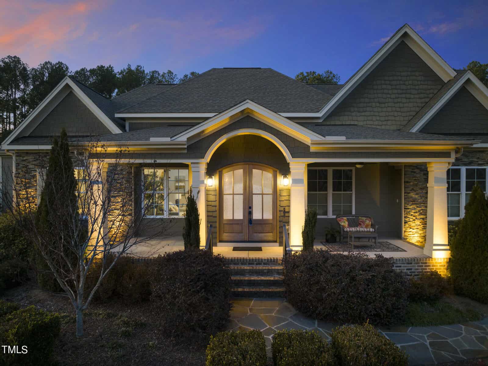 Front view of a large house with stone and wood accents, illuminated by warm exterior lighting in the evening. A covered porch with columns and a cozy seating area is visible, surrounded by well-maintained landscaping.