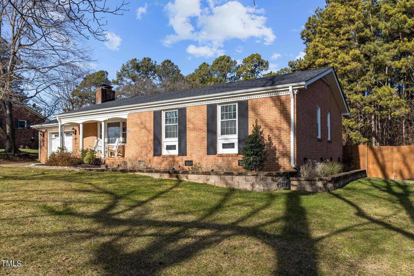 A single-story brick house on 500 Sunset Avenue in Oxford features white trim and a covered front porch with rocking chairs. The yard is grassy, with a few trees casting long shadows. Pine trees stand in the background under a blue sky with scattered clouds.