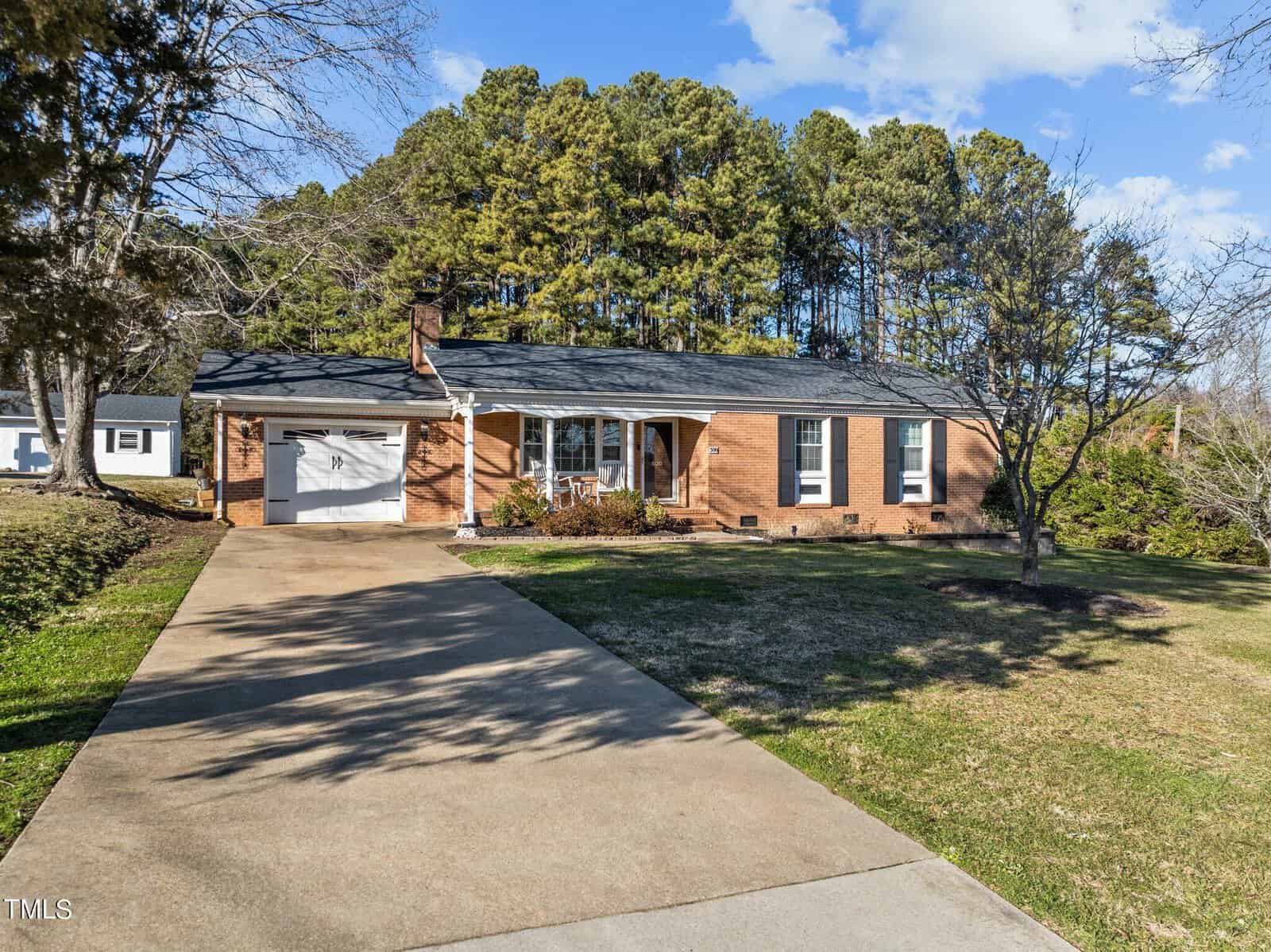 A single-story brick house with a gray roof, a garage, and a concrete driveway. There are trees behind the house and a lawn with sparse grass in front. The sky is partly cloudy.