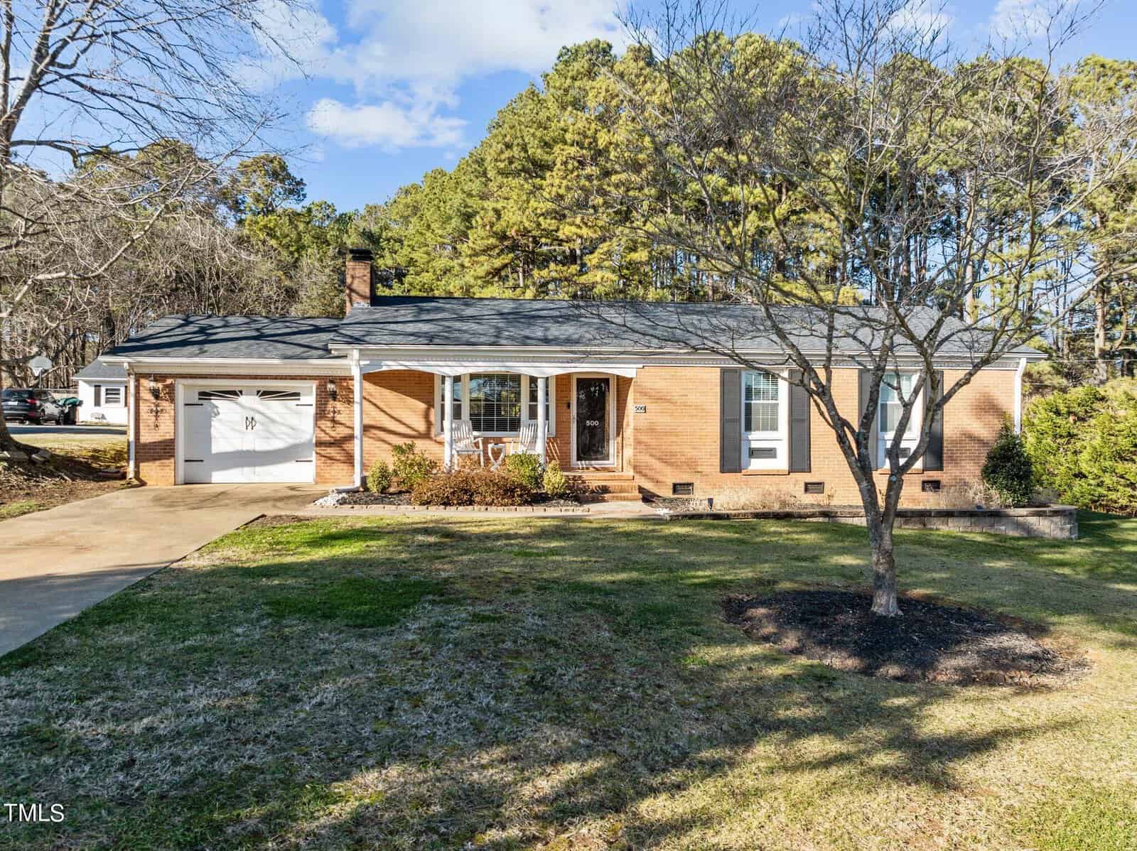 A single-story brick house with a black roof stands in a grassy yard. It has a white door and large windows. A driveway leads to an attached garage. The background features tall trees and a clear blue sky.