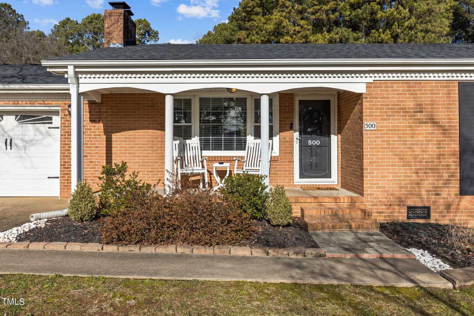 A brick house with a covered front porch featuring two white rocking chairs and a small table. The house number "500" is displayed on the door and wall. The front yard has shrubs and a paved walkway. A garage door is partially visible on the left.