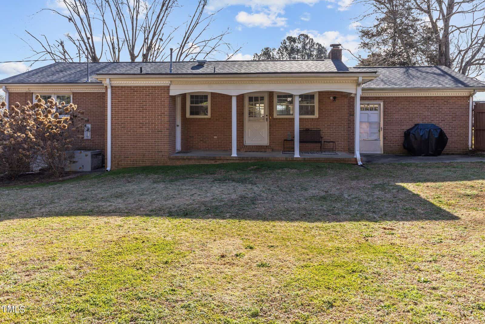 A brick house with a covered back patio, featuring a few outdoor chairs. There's a grassy backyard, leafless trees in the background, and a grill on the right side of the house. The sky is partly cloudy.