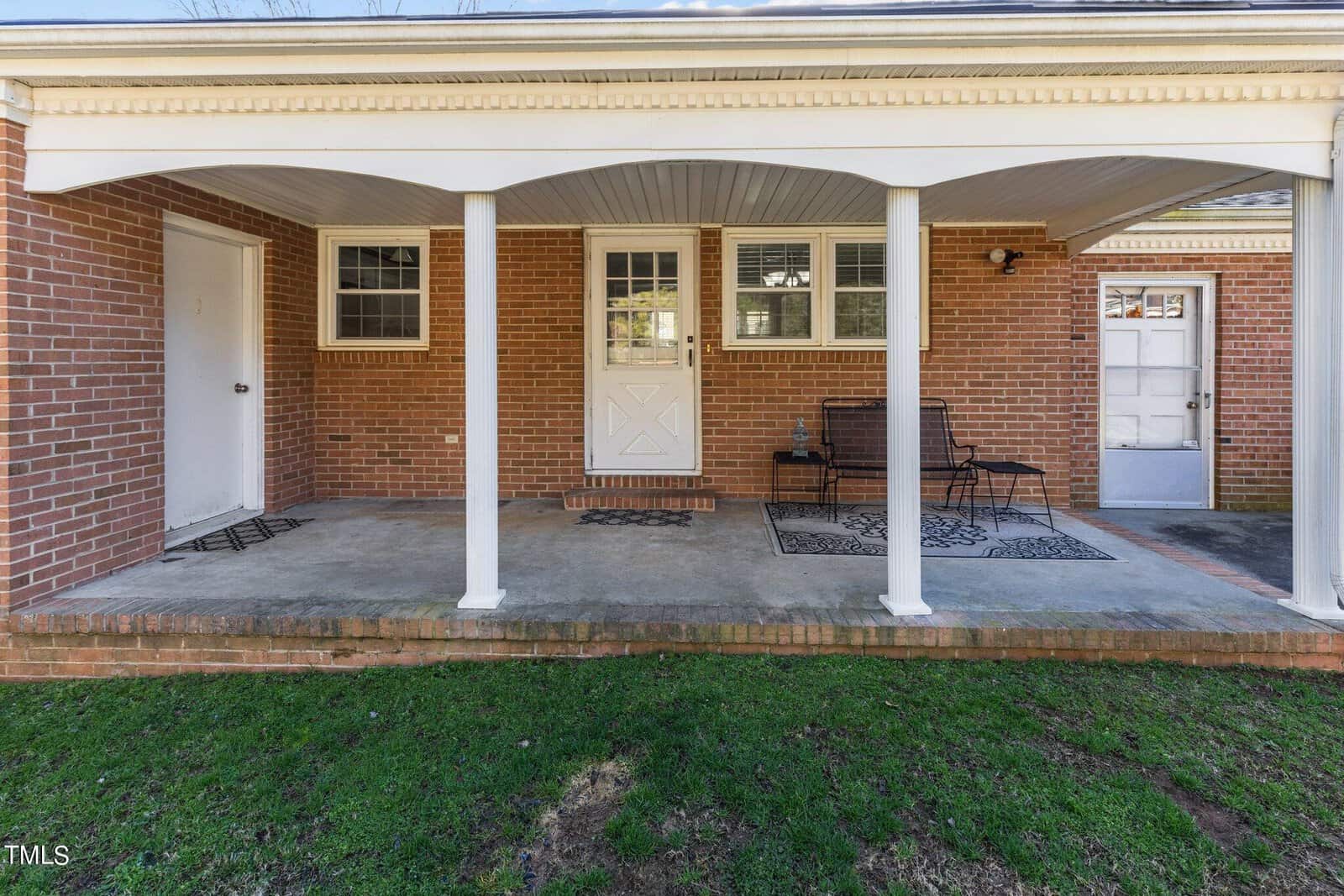 A brick house features a covered porch with a concrete floor. The porch has a white ceiling and columns. Two doors and three windows are visible. A black metal bench and two decorative rugs enhance the space. Grass grows in front of the porch.